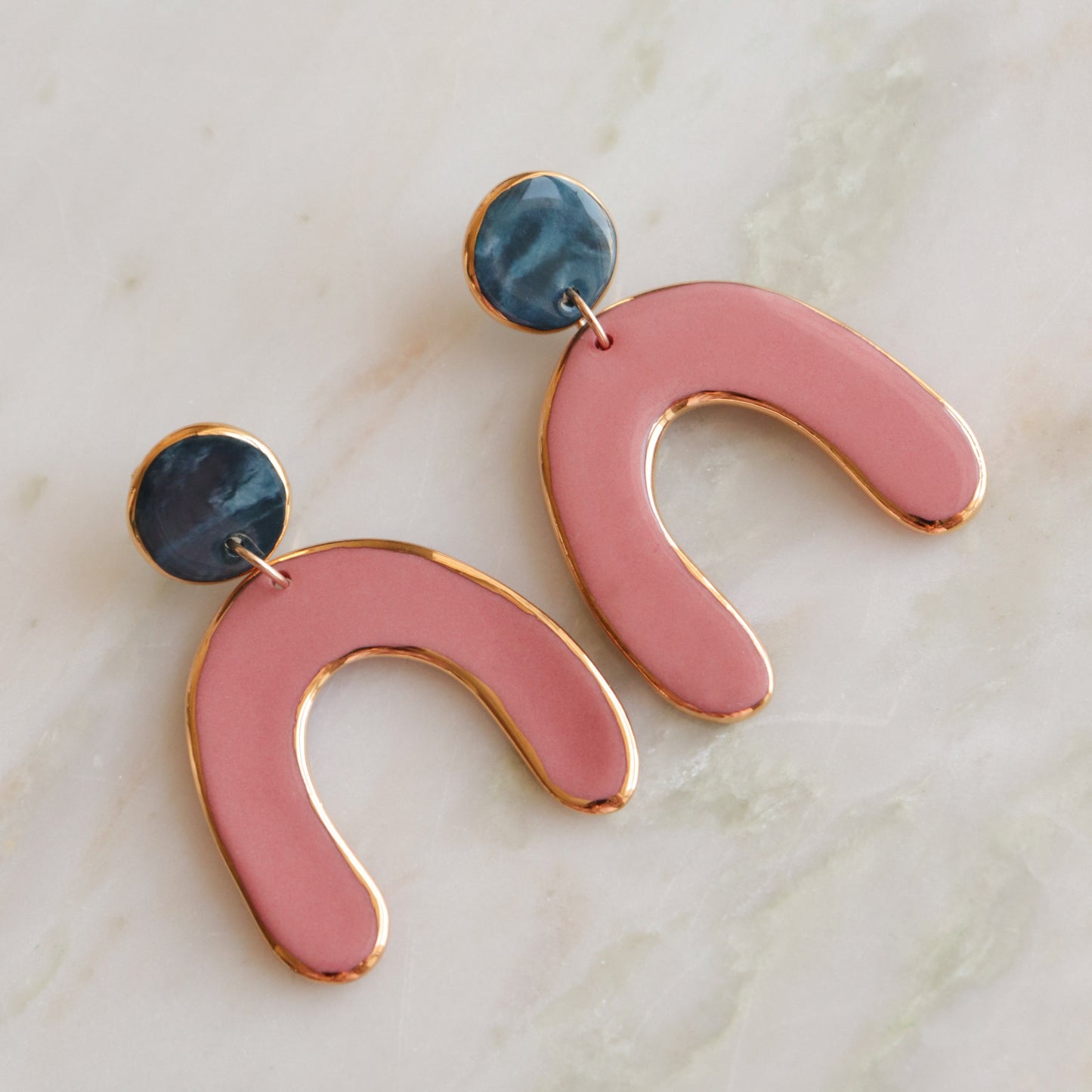 The Arquus Earrings by Rozenthal Jewelry feature porcelain statement pieces with blue circular studs and pink, 24k gold-edged U-shaped dangles, shown on a light marble surface.