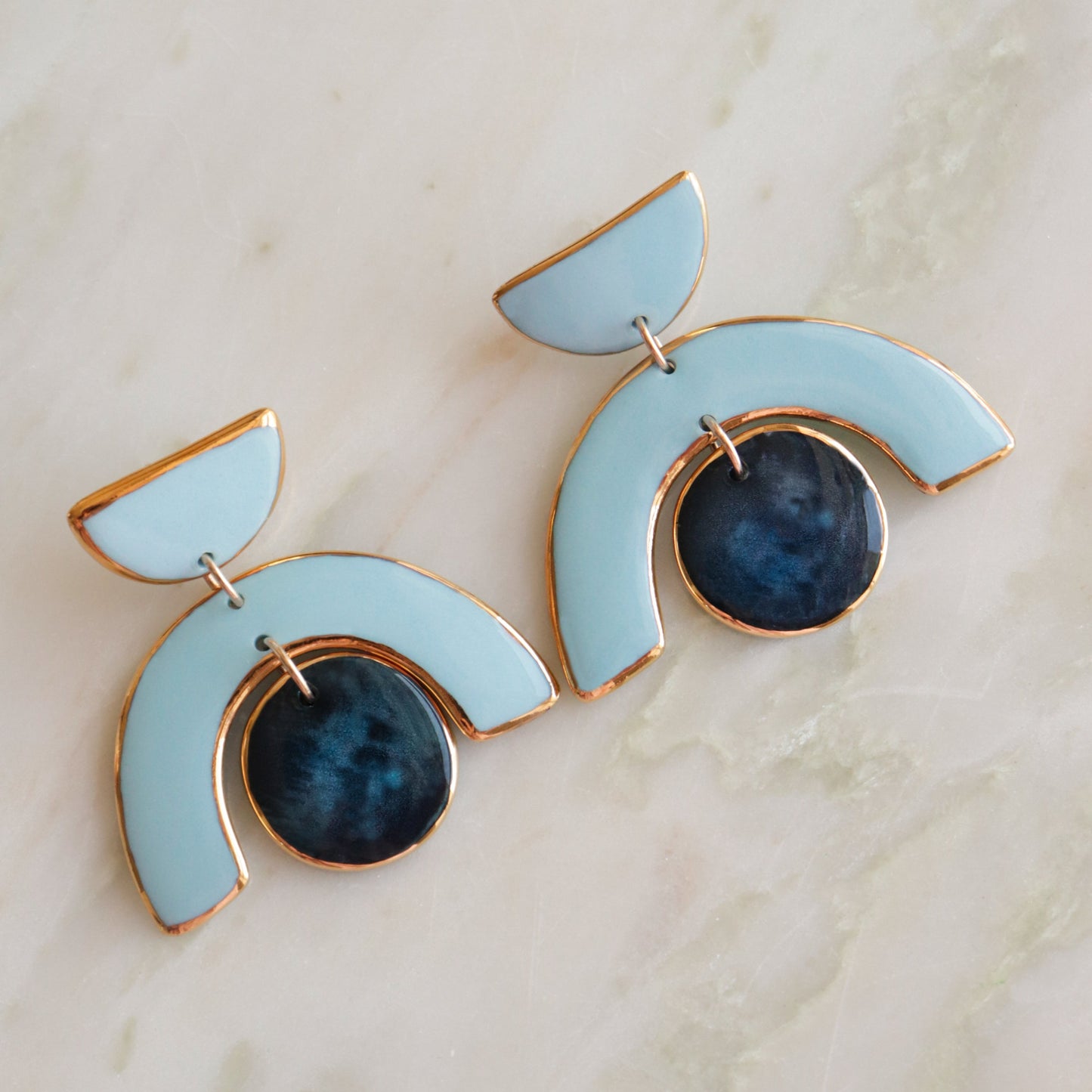 The Arquus Earrings by Rozenthal Jewelry are modern porcelain statement earrings with light blue semi-circles outlined in 14k gold-filled accents and dark blue circular pendants, shown on a light marble surface.