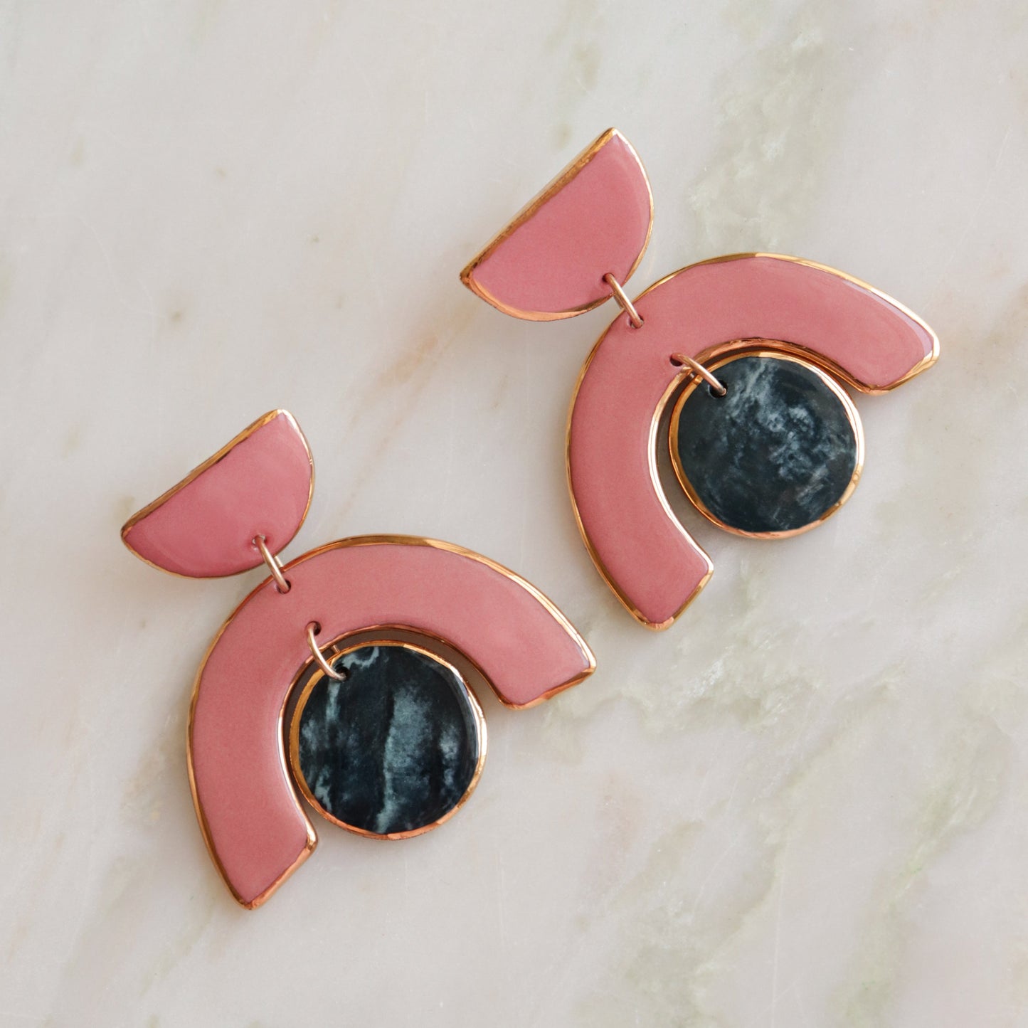 The Arquus Earrings by Rozenthal Jewelry feature porcelain pink, gold-edged arches with dark marbled pendants and 14k gold-filled accents, beautifully displayed on a light marble surface.