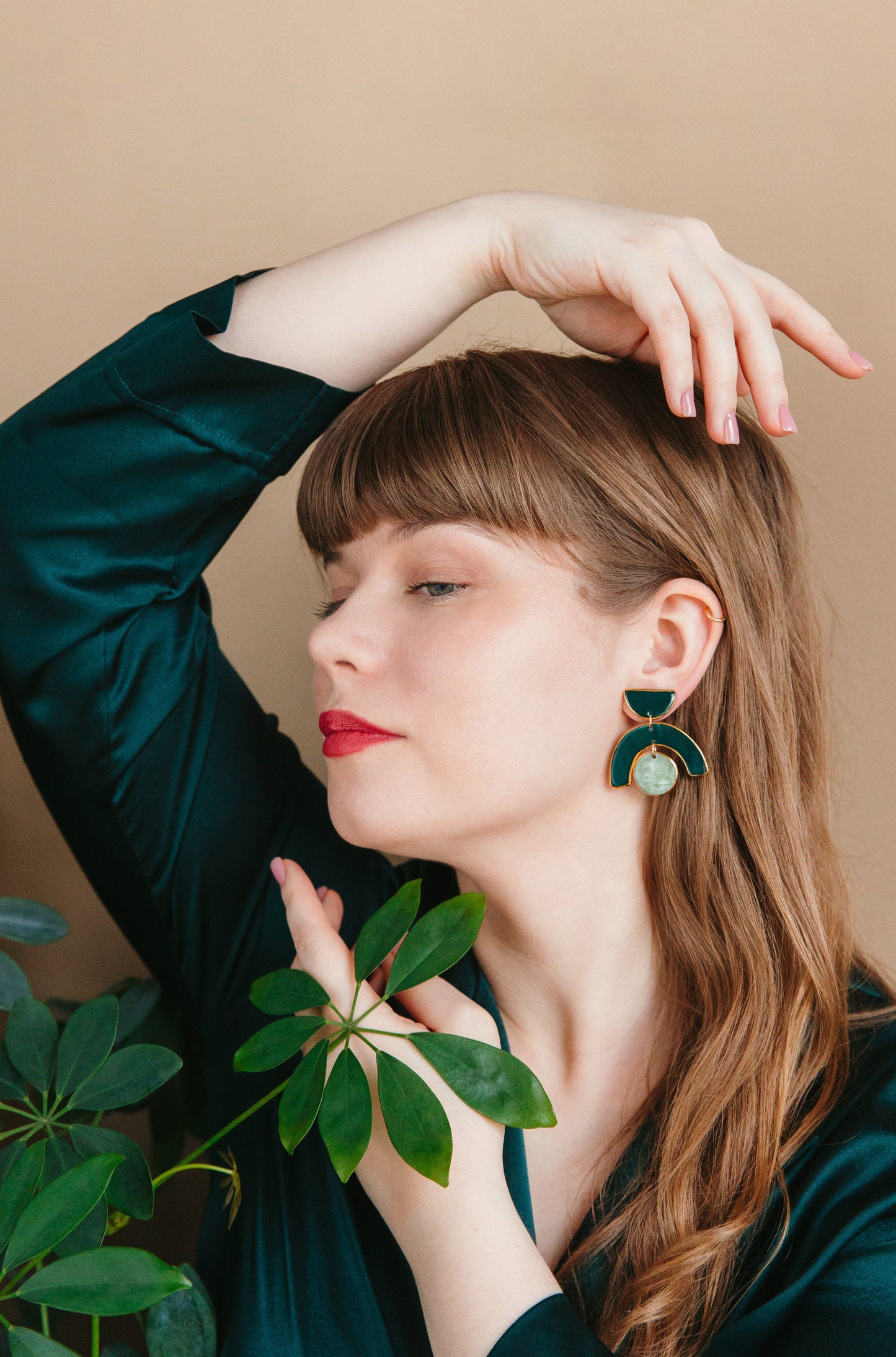 A woman with long brown hair and closed eyes poses gracefully, holding a leafy green plant near her face. She wears Rozenthal Jewelrys Arquus Earrings and a dark green top, set against a beige background.