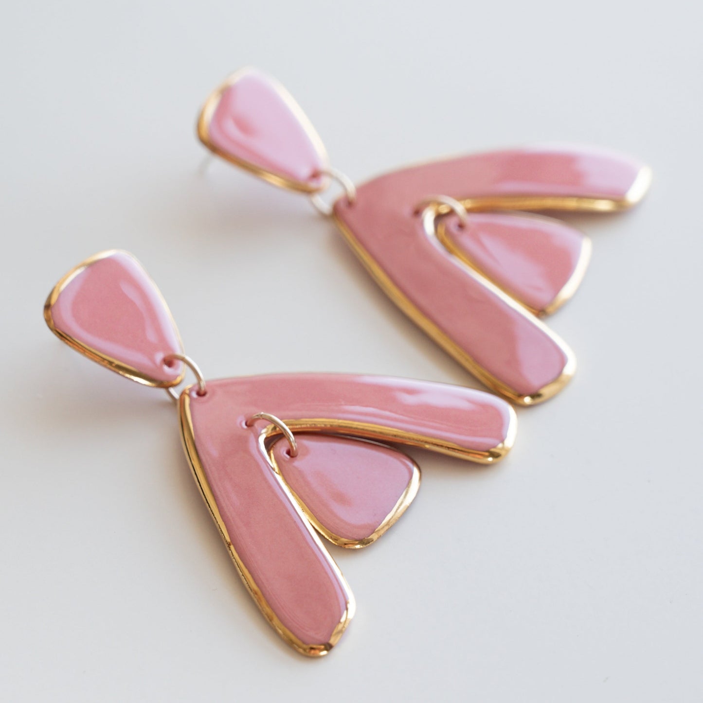 The Arquus Earrings by Rozenthal Jewelry are pink porcelain statement earrings with 24K gold edging and abstract, fan-shaped designs, shown on a white background.