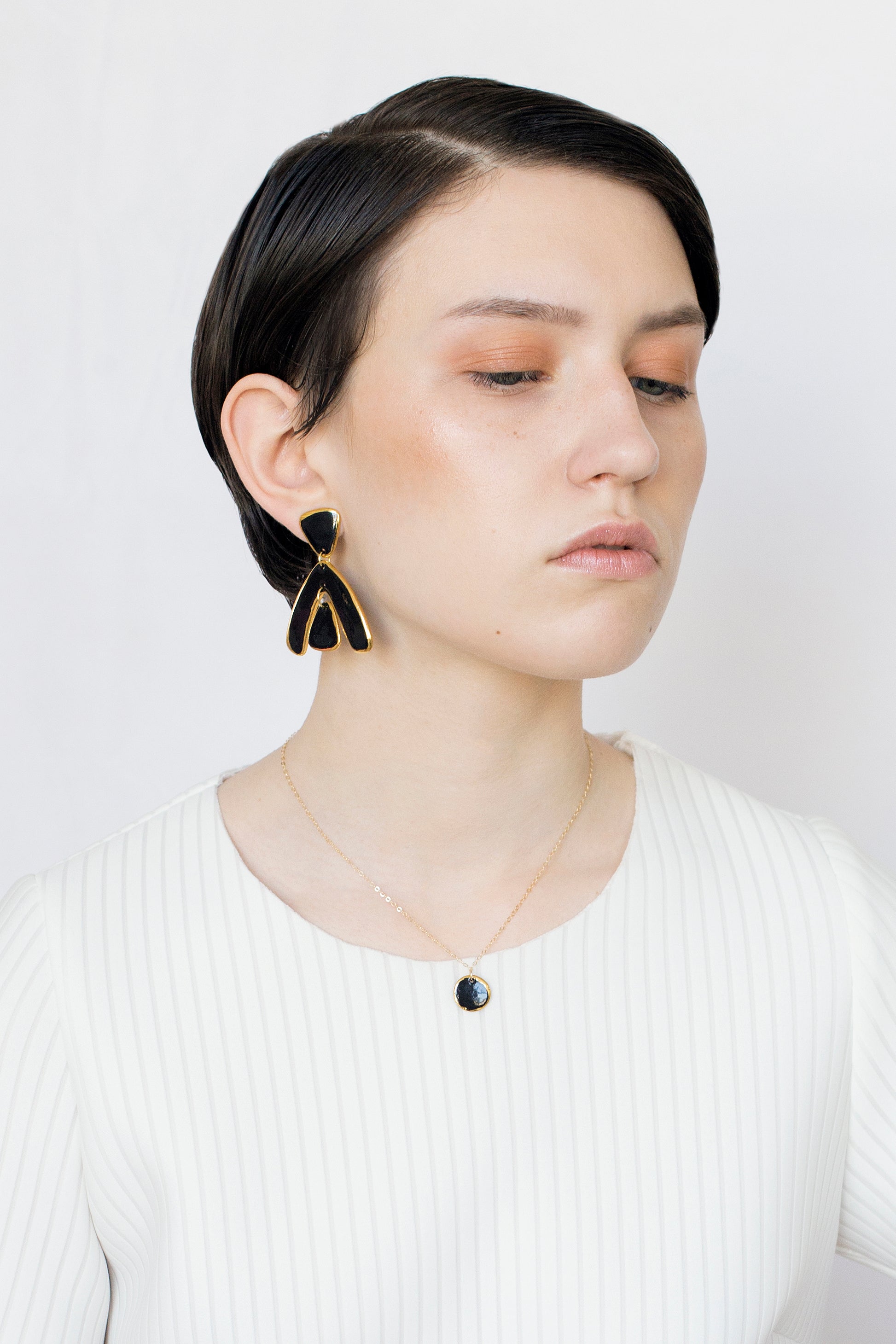 A woman with short dark hair wears a white ribbed top, Rozenthal Jewelry’s Arquus Earrings in black and gold, and a delicate gold necklace with a dark pendant, looking down with a neutral expression against a light background.