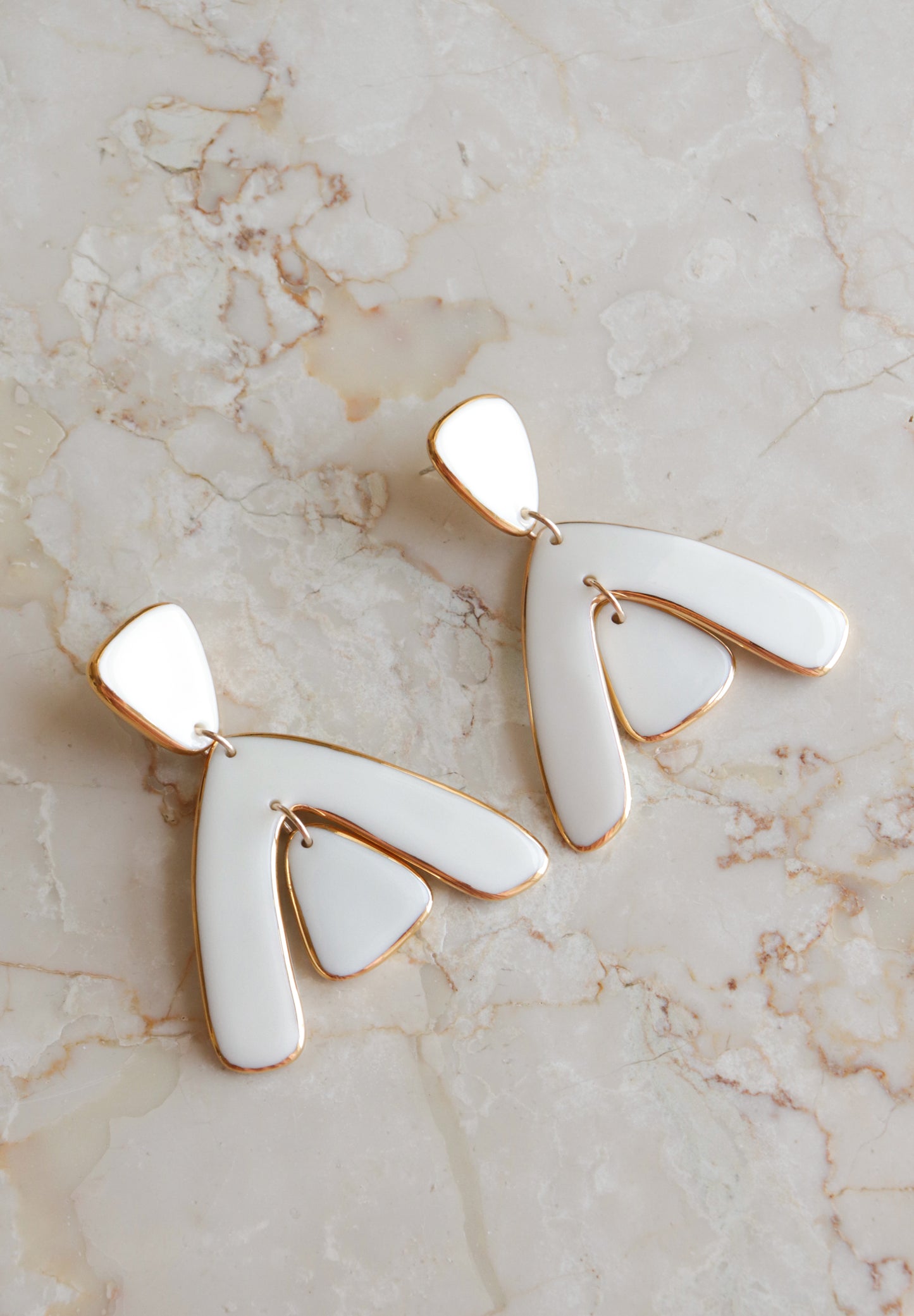 The Arquus Earrings by Rozenthal Jewelry are modern abstract porcelain statement pieces with white enamel and 24K gold trim, featuring three curved overlapping segments, displayed on a light marble surface.