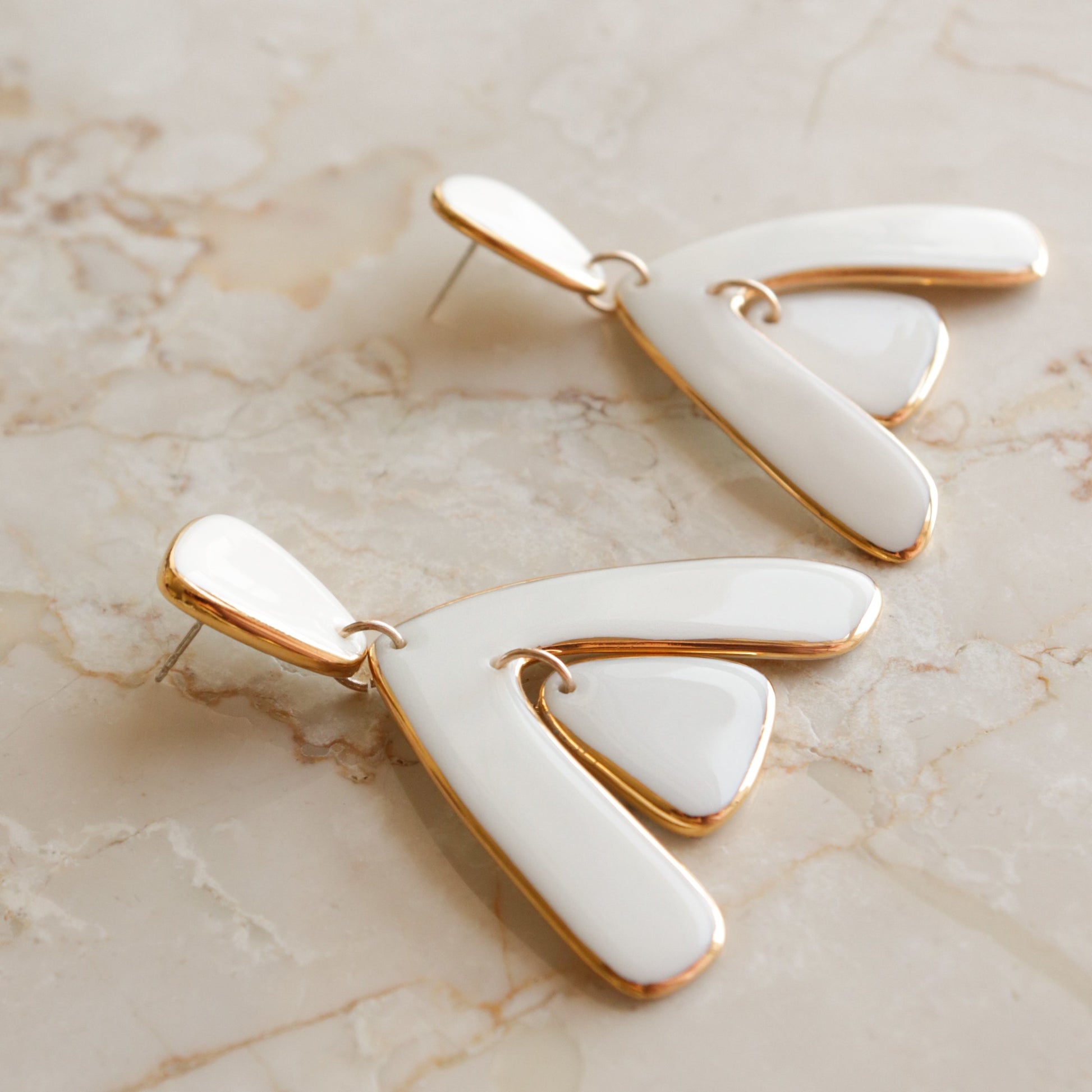 The Arquus Earrings by Rozenthal Jewelry are modern, abstract porcelain statement pieces with 24K gold edges. Each features three curved shapes radiating from a central point, and is displayed on a light marble surface.