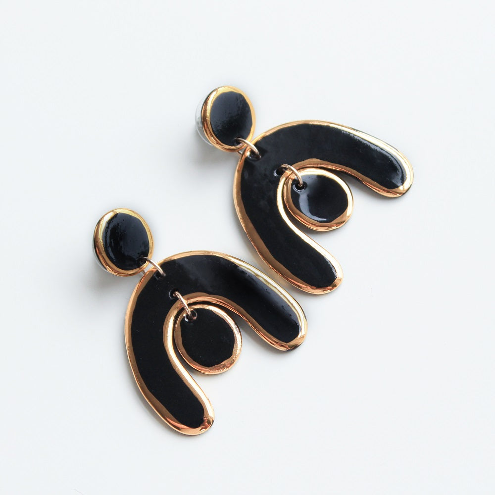 The Arquus Earrings by Rozenthal Jewelry are porcelain statement earrings with abstract black and 14k gold-filled details, featuring circular studs and curved dangles with a center circle, shown on a white background.