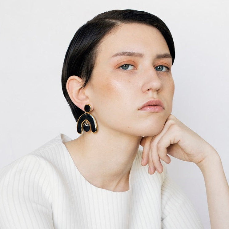 A person with short dark hair, wearing a white textured top and Rozenthal Jewelrys Arquus Earrings—porcelain statement pieces with 14k gold-filled accents—rests their chin on their hand, gazing thoughtfully at the camera against a plain light background.