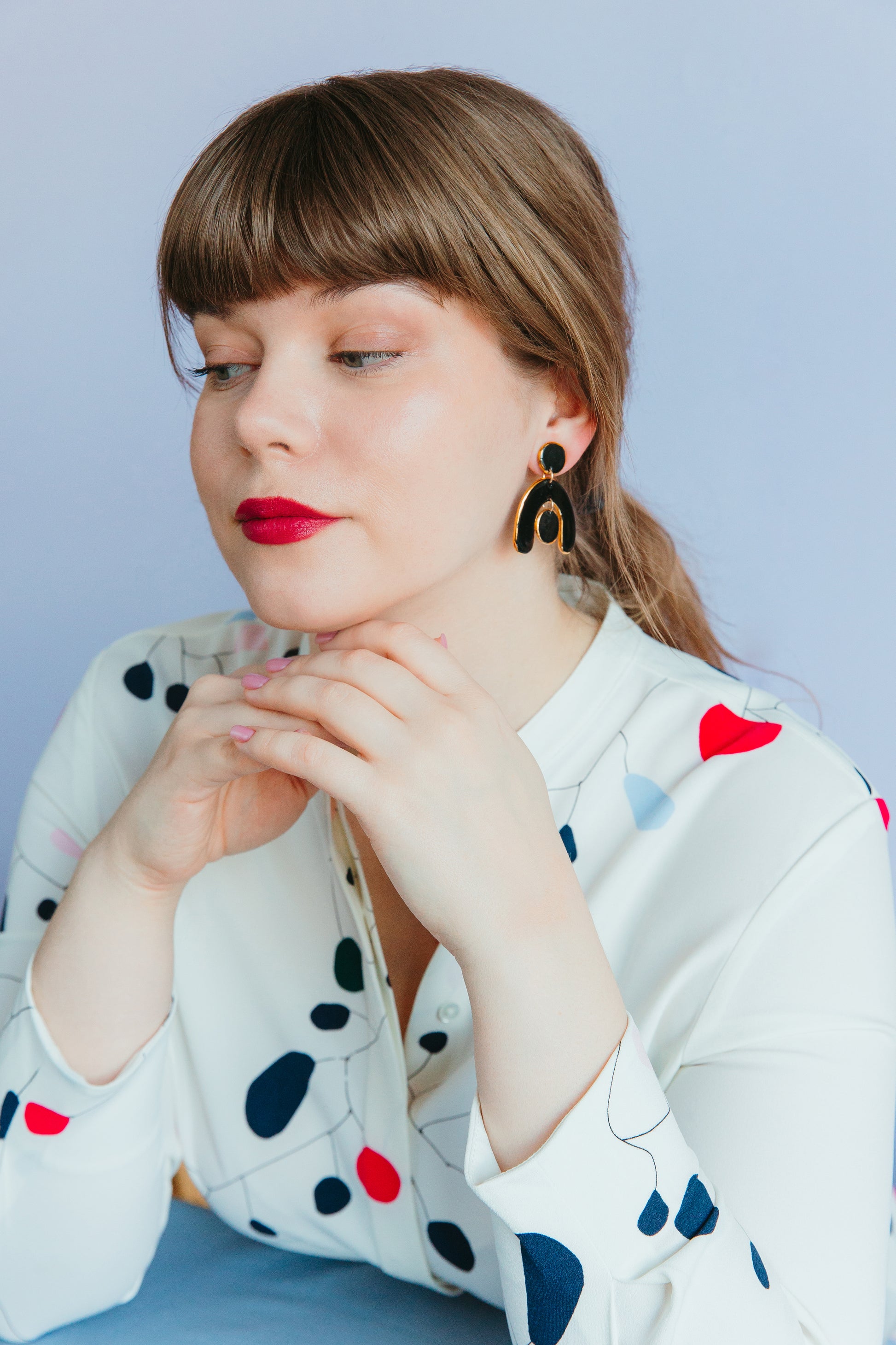A woman with light brown hair and red lipstick wears Rozenthal Jewelrys Arquus Earrings and a white blouse with colorful abstract patterns, resting her chin on her hands as she looks to the side against a pale purple background.