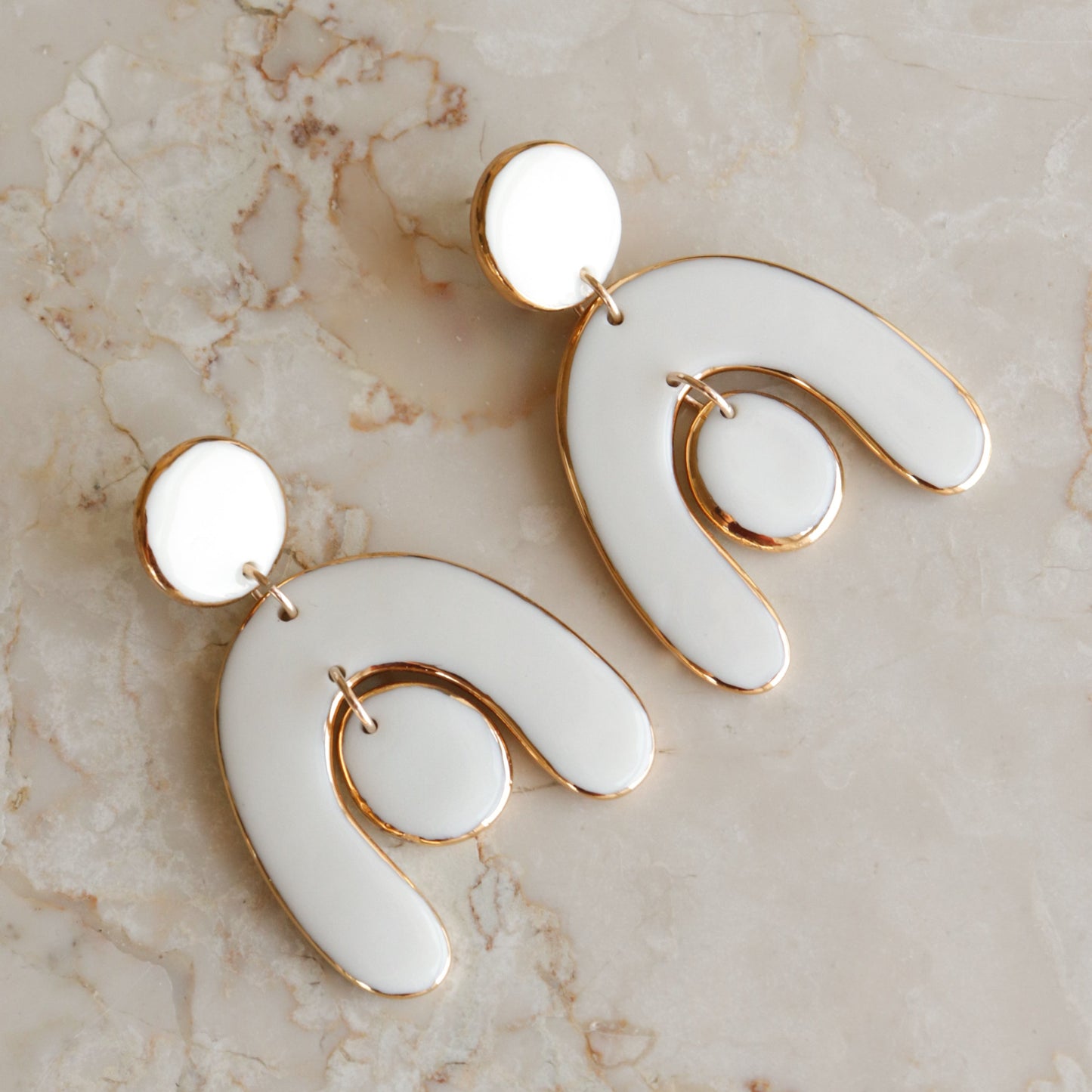 The Arquus Earrings by Rozenthal Jewelry feature modern, abstract porcelain with 14k gold-filled trim, handmade in a curved arch shape with a central circular accent, displayed on a light beige marble surface.