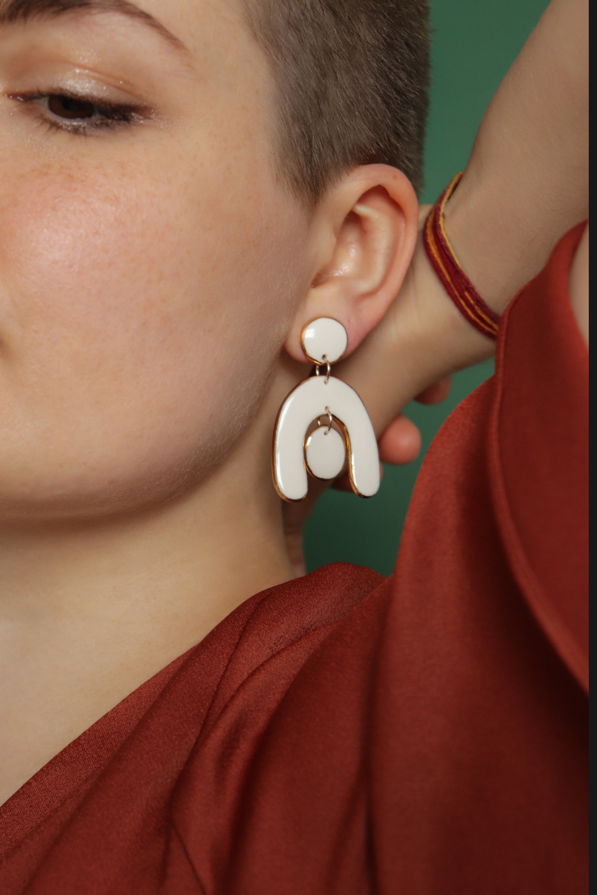 A person with short hair wears a rust top and the Arquus Earrings by Rozenthal Jewelry—large white porcelain statement pieces. Their hand, with a colorful string bracelet, is raised to their head against a green background.