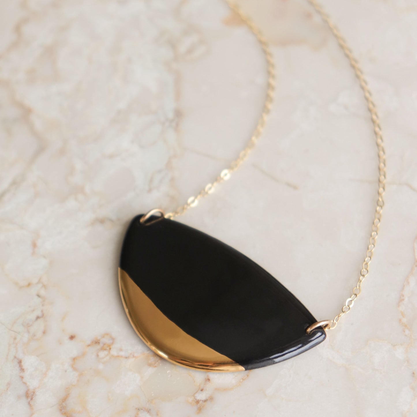 The Leaf Necklace by Rozenthal Jewelry features a semicircular pendant, half black and half gold, on a gold chain—an eye-catching handmade piece ideal for any minimalist jewelry collection. Displayed on a light marble surface.