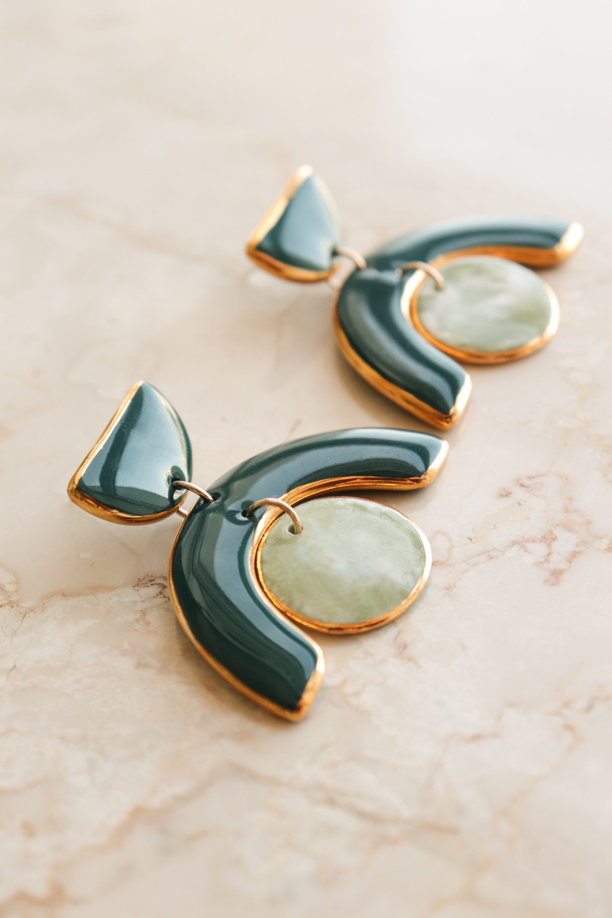 The Arquus Earrings by Rozenthal Jewelry are modern porcelain statement earrings featuring curved green enamel edged in 14k gold-filled detail and light green marble-like pendants, shown on a light marble surface.