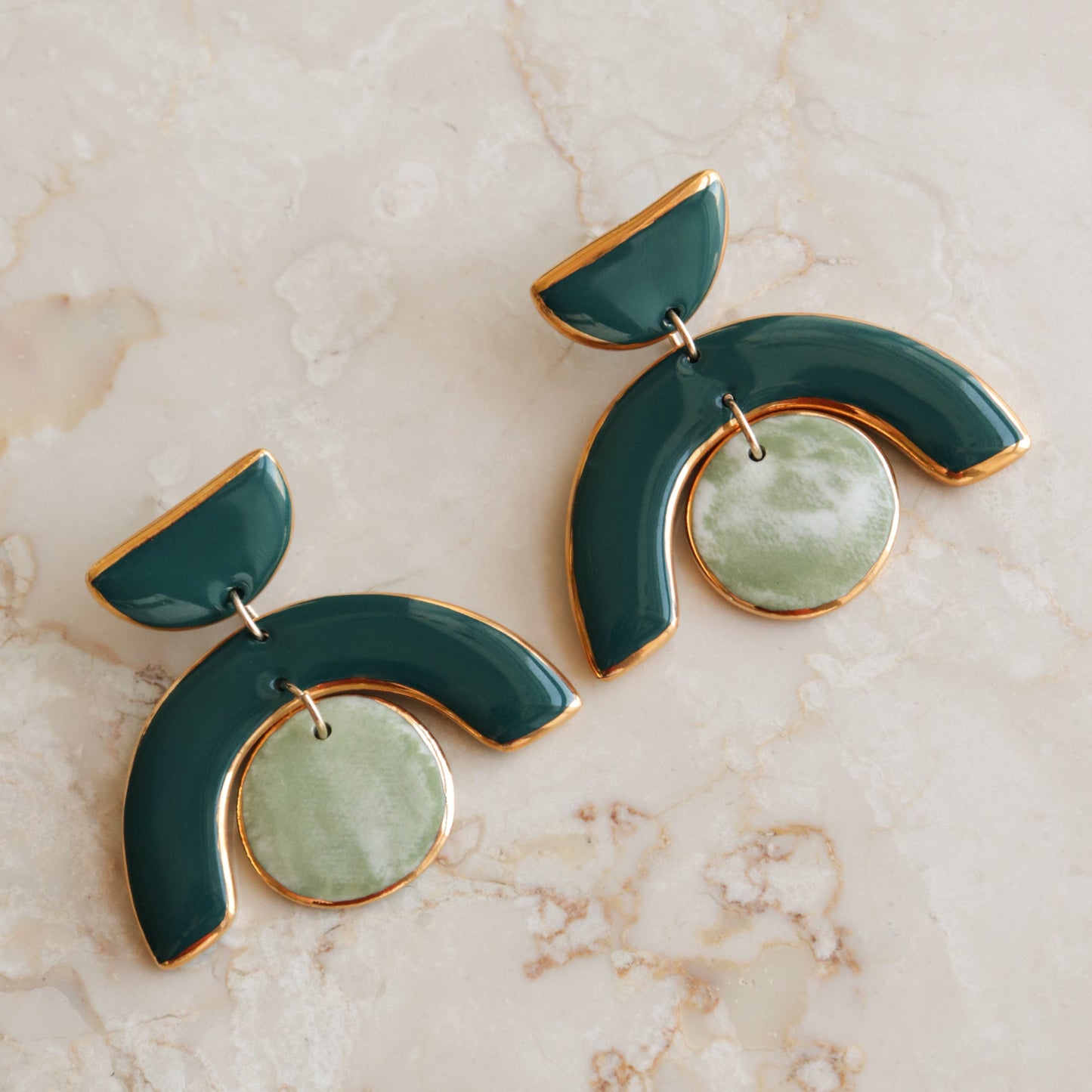 The Arquus Earrings by Rozenthal Jewelry are modern green and gold porcelain statement pieces featuring semi-circular shapes and round marbled green pendants, shown on a light marble surface.
