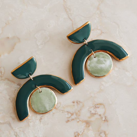 The Arquus Earrings by Rozenthal Jewelry are modern green and gold porcelain statement pieces featuring semi-circular shapes and round marbled green pendants, shown on a light marble surface.