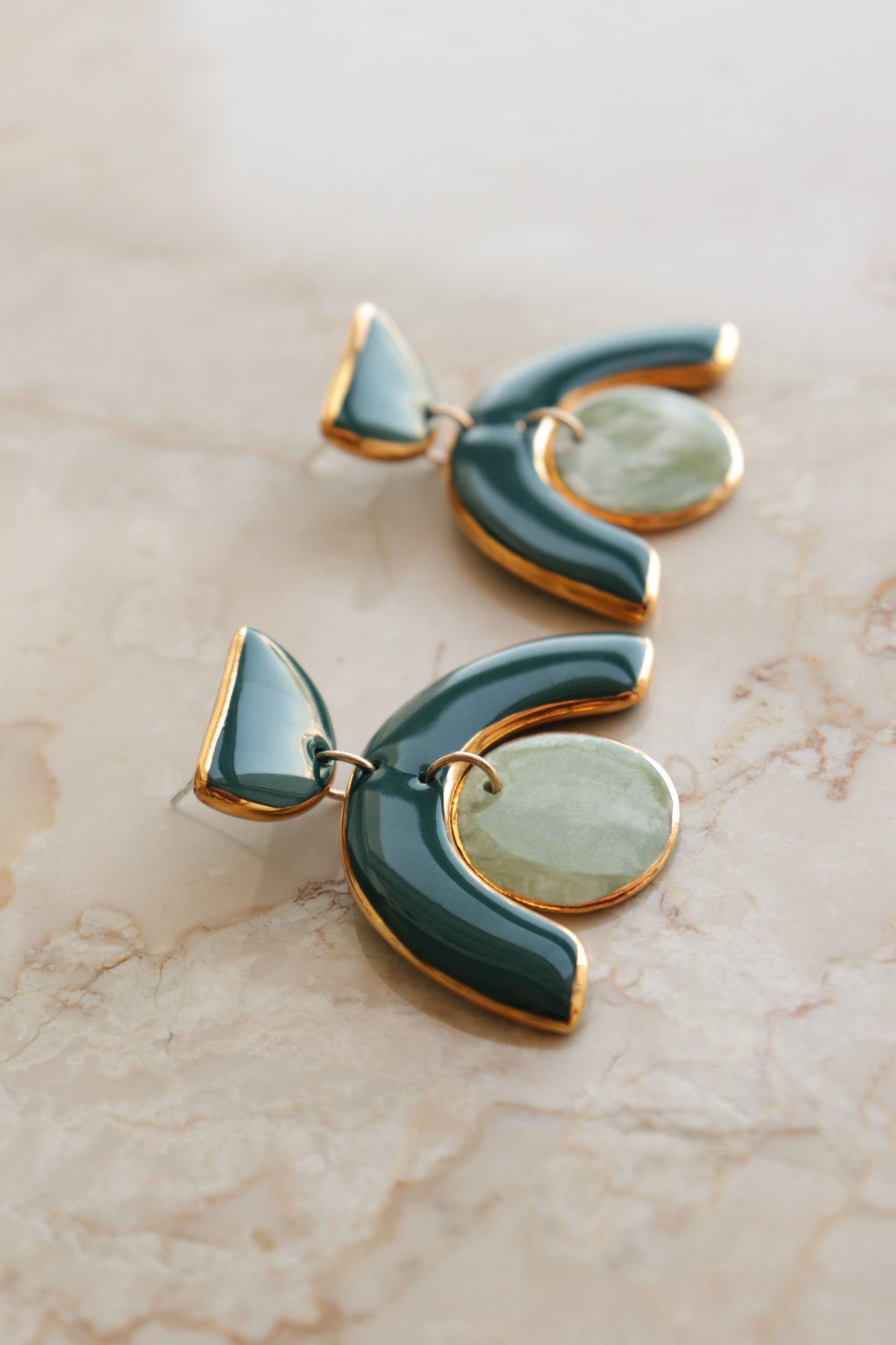 The Arquus Earrings by Rozenthal Jewelry are modern, abstract porcelain statement earrings with 14k gold-filled trim, teal curves, and round light green stone accents, shown on a marble surface.