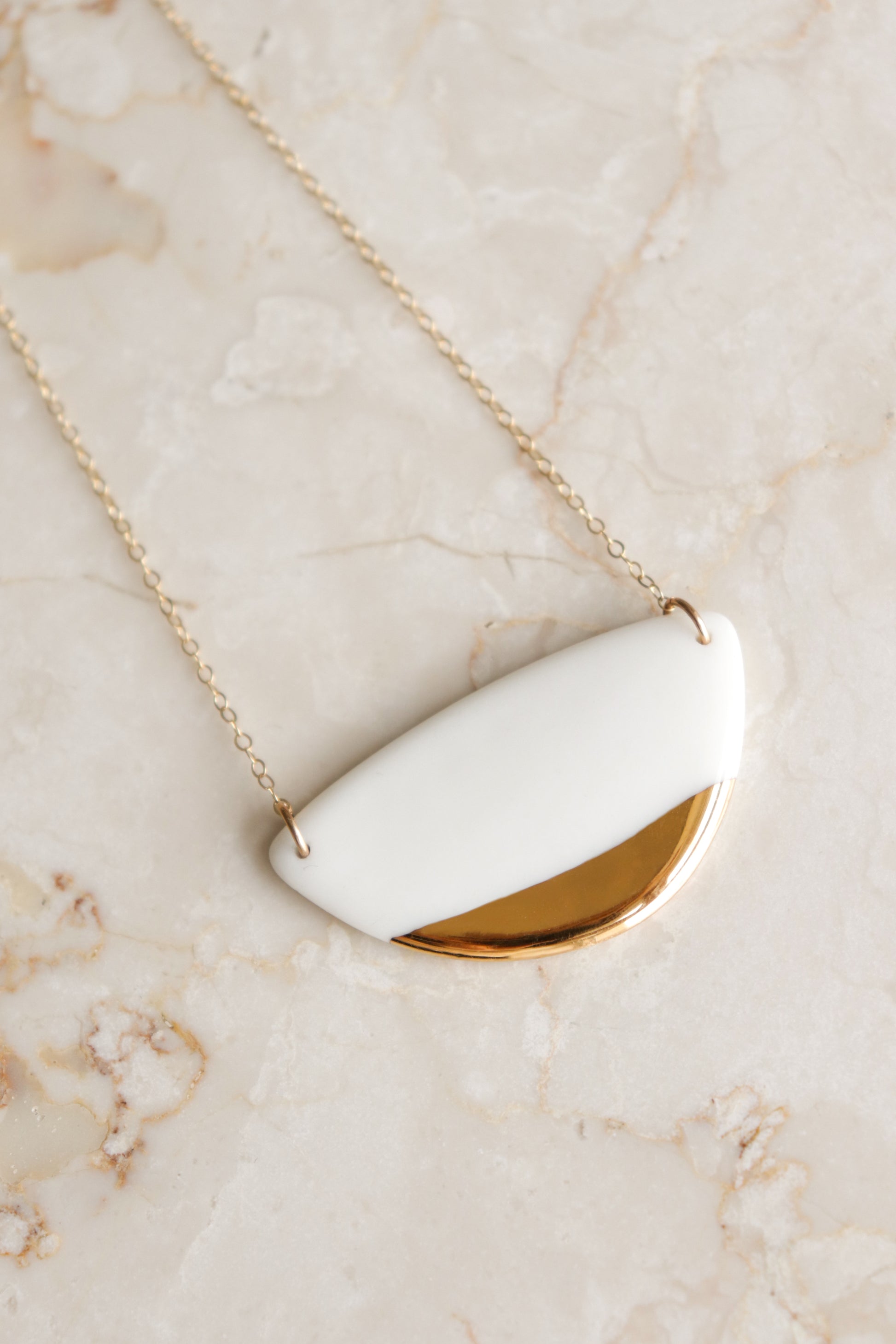 The Leaf Necklace by Rozenthal Jewelry features a white and gold semi-circular porcelain pendant on a gold chain, elegantly displayed on a light marble surface—a refined piece of minimalist jewelry.
