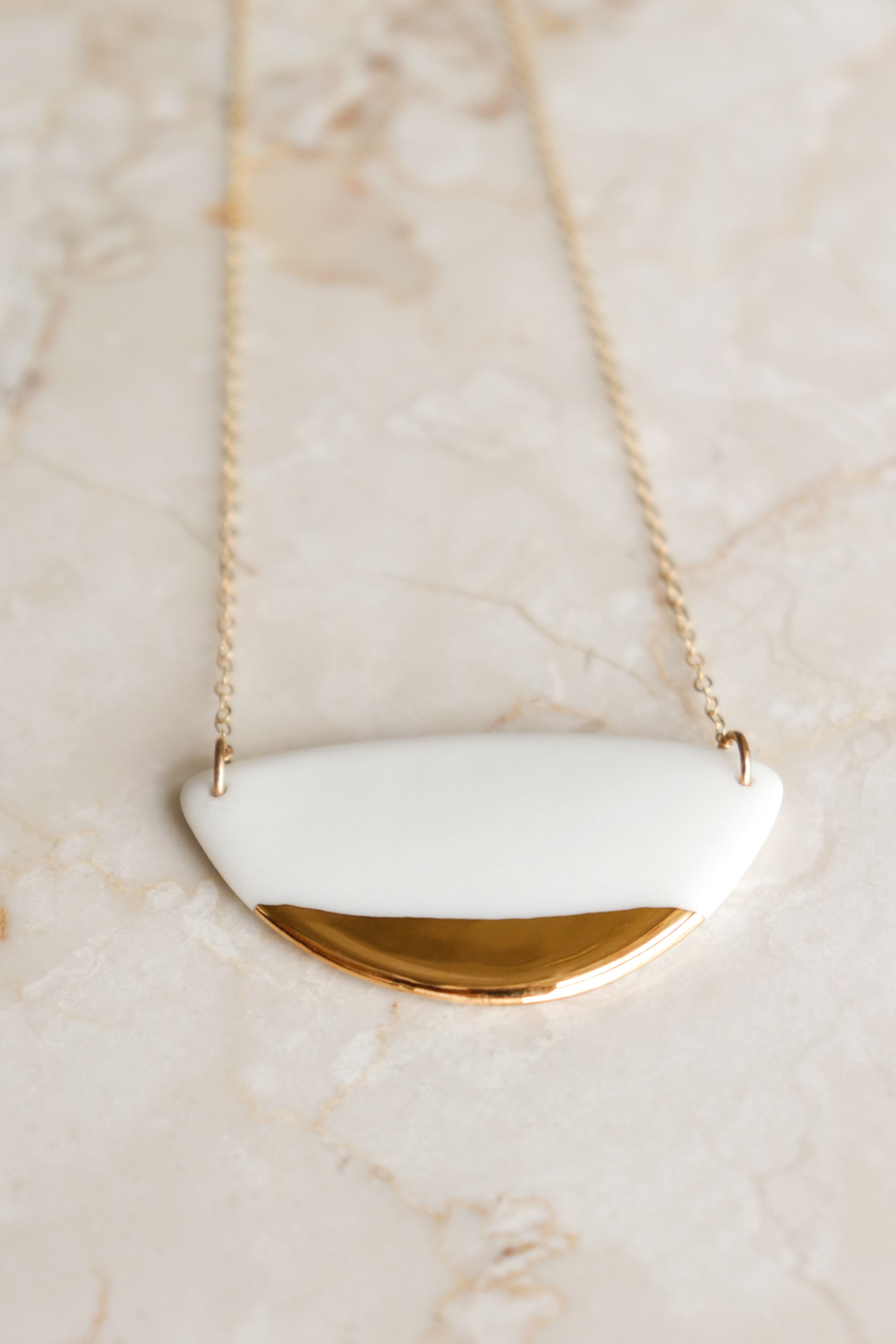 The Leaf Necklace by Rozenthal Jewelry features a gold chain and a white semi-circular pendant with a shiny gold accent along the edge, offering minimalist elegance. Displayed on a light marble surface.