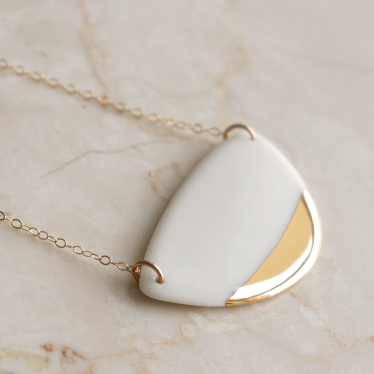 The Leaf Necklace by Rozenthal Jewelry features a sleek gold chain with a modern pendant—a smooth, white semi-circular ceramic piece accented by shiny gold—showcasing minimalist elegance on a light marble background.