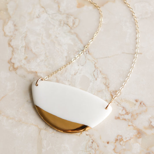 The Leaf Necklace by Rozenthal Jewelry is a minimalist gold chain with a pendant of half-white, half-gold semicircles, shown on a light marble surface.