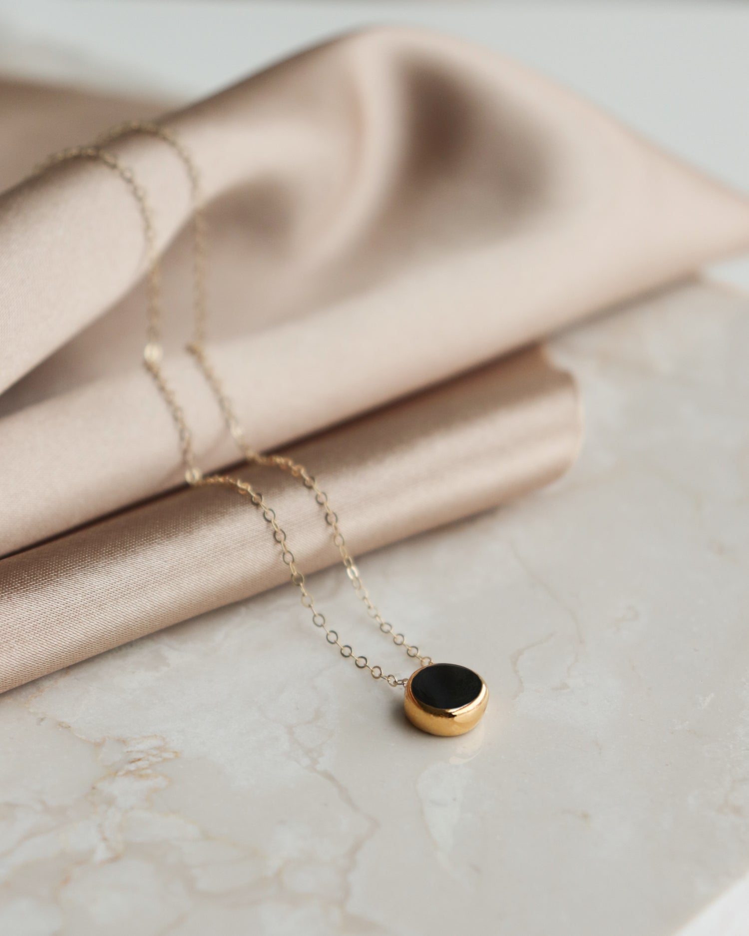 The Round Necklace by Rozenthal Jewelry, featuring a minimalist gold chain and round black pendant, is displayed on marble beside draped beige satin.