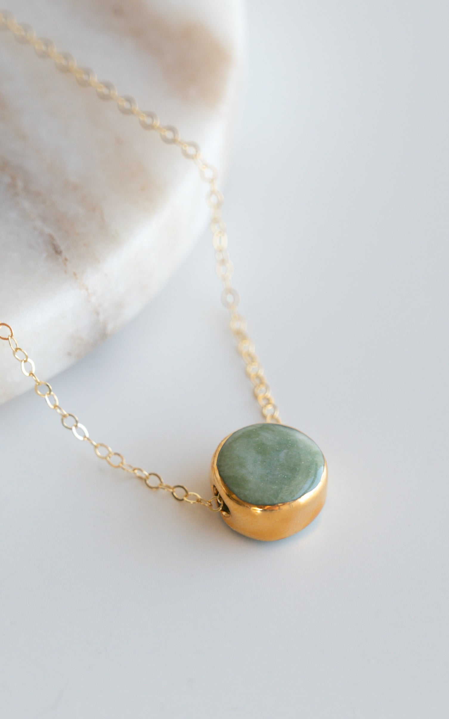The Round Necklace by Rozenthal Jewelry features a gold chain with a round green gemstone pendant in a gold bezel, displayed as minimalist jewelry on a white and beige marble surface.