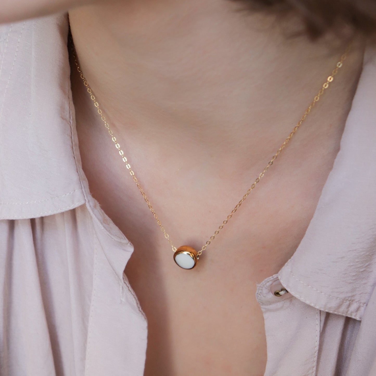 A close-up of the Rozenthal Jewelry Round Necklace—a minimalist gold chain with a small round white porcelain pendant—worn against light skin and a pale pink collared shirt.