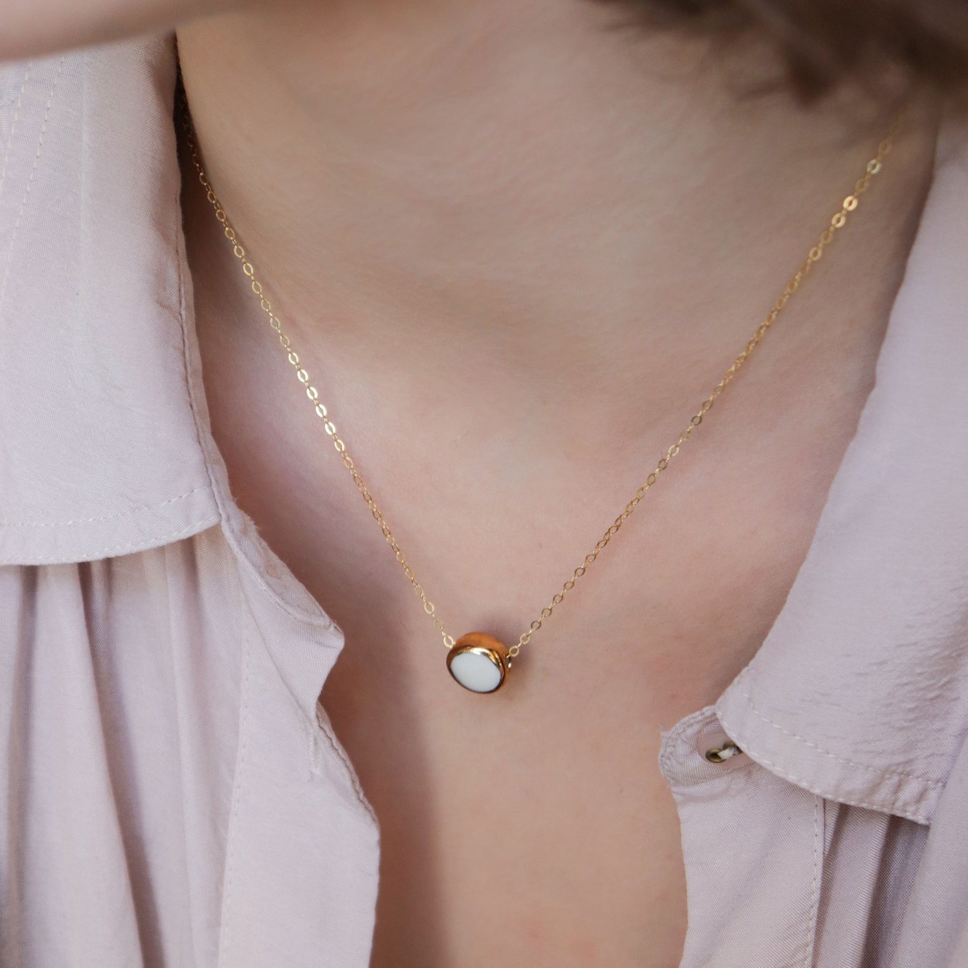 A close-up of the Rozenthal Jewelry Round Necklace—a minimalist gold chain with a small round white porcelain pendant—worn against light skin and a pale pink collared shirt.