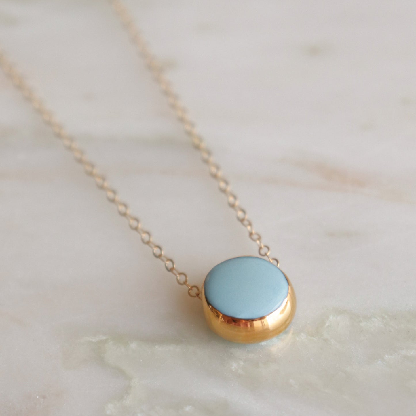 The Rozenthal Jewelry Round Necklace features a delicate gold chain with a round, light blue stone pendant in a gold bezel, highlighting minimalist style and displayed on soft marble.