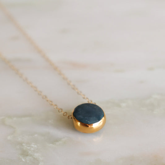 The Round Necklace by Rozenthal Jewelry features a delicate gold chain with a round, dark blue stone pendant—a stunning minimalist piece ideal for handmade jewelry lovers, beautifully displayed on a marbled surface.