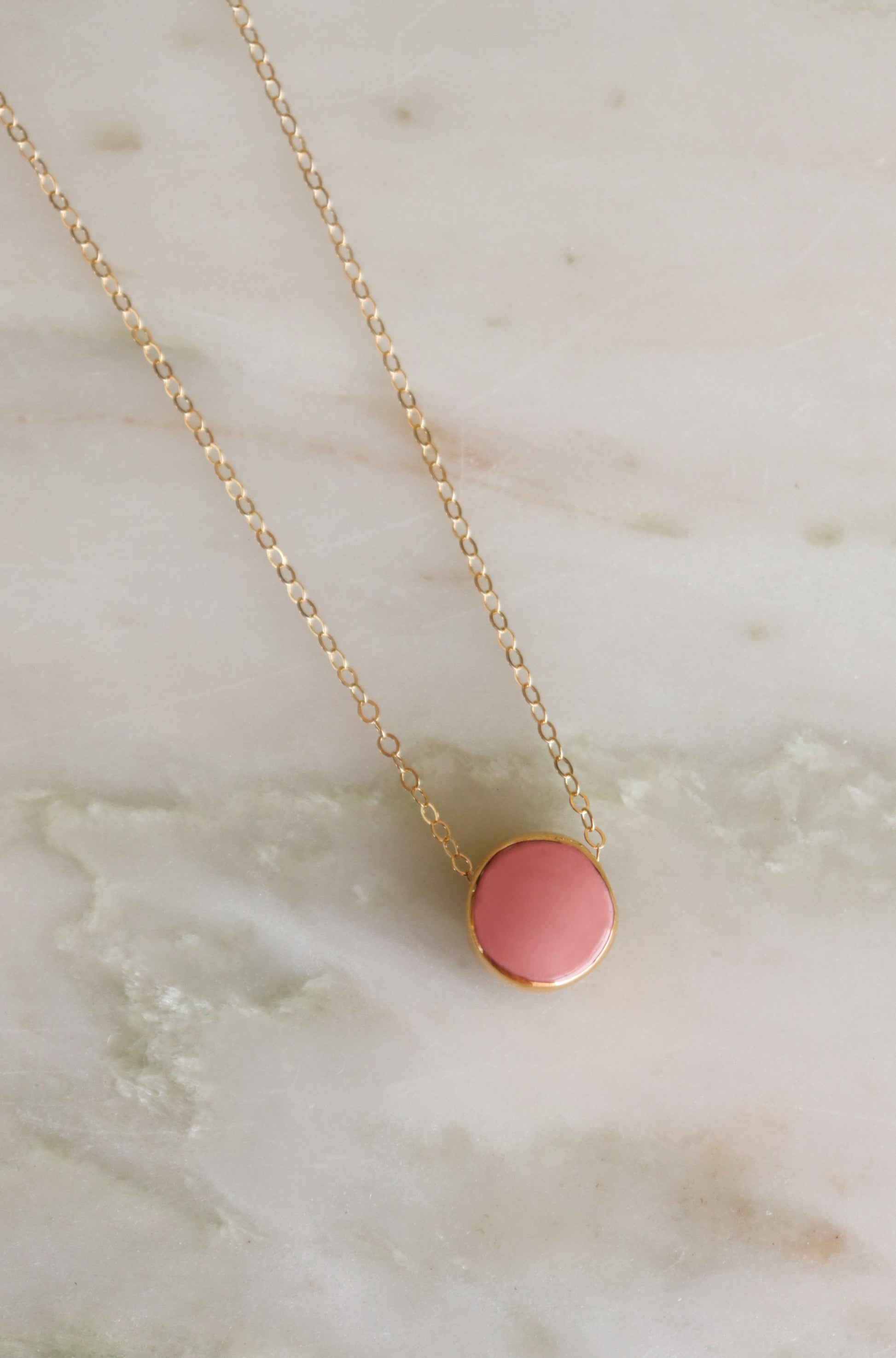 The Rozenthal Jewelry Round Necklace features a gold chain and a round pink porcelain pendant, shown resting on light marble with green and beige veining for a minimalist look.