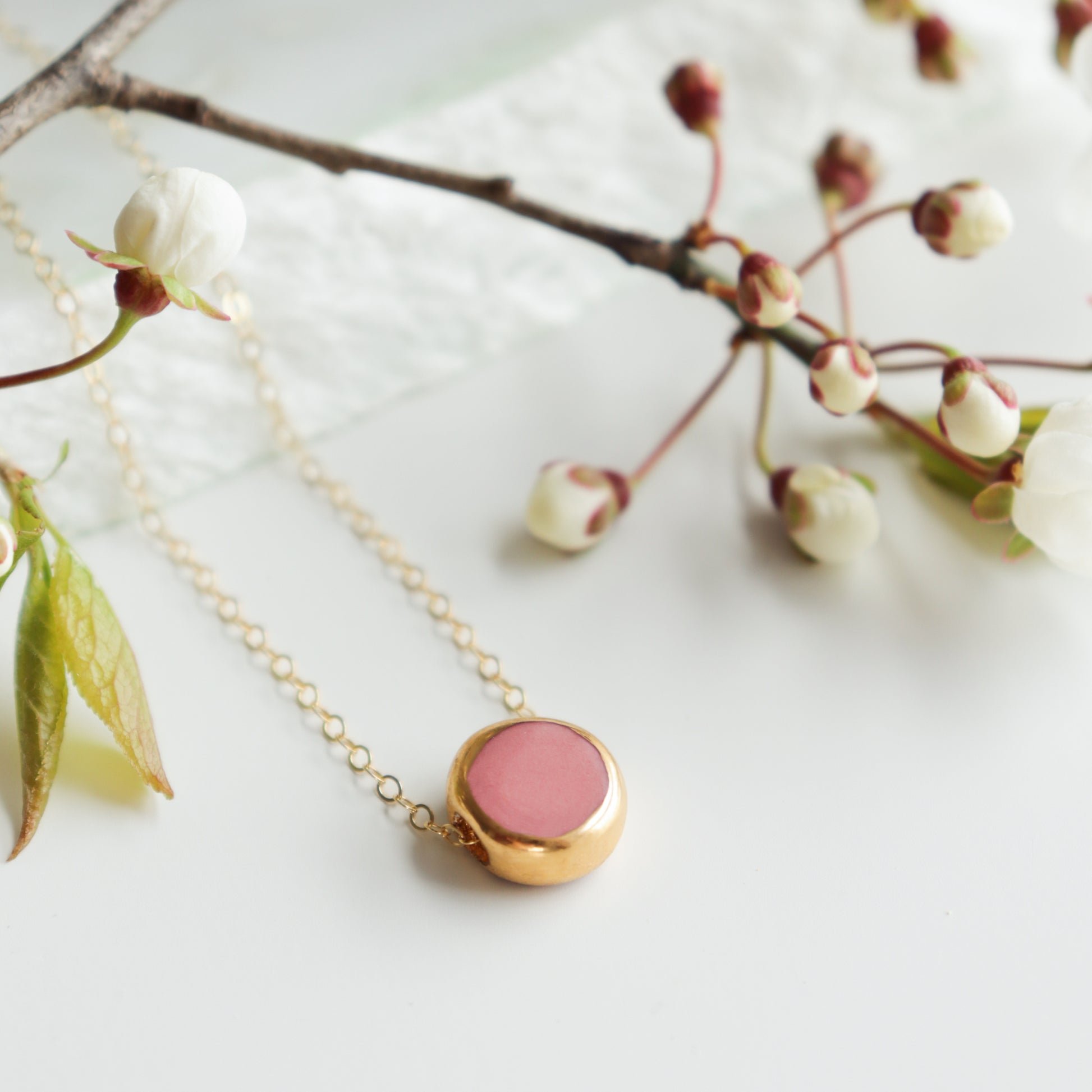 The Rozenthal Jewelry Round Necklace, featuring a round pink pendant and ideal for minimalist jewelry fans, is displayed on a white surface beside delicate white blossom flowers and green leaves.