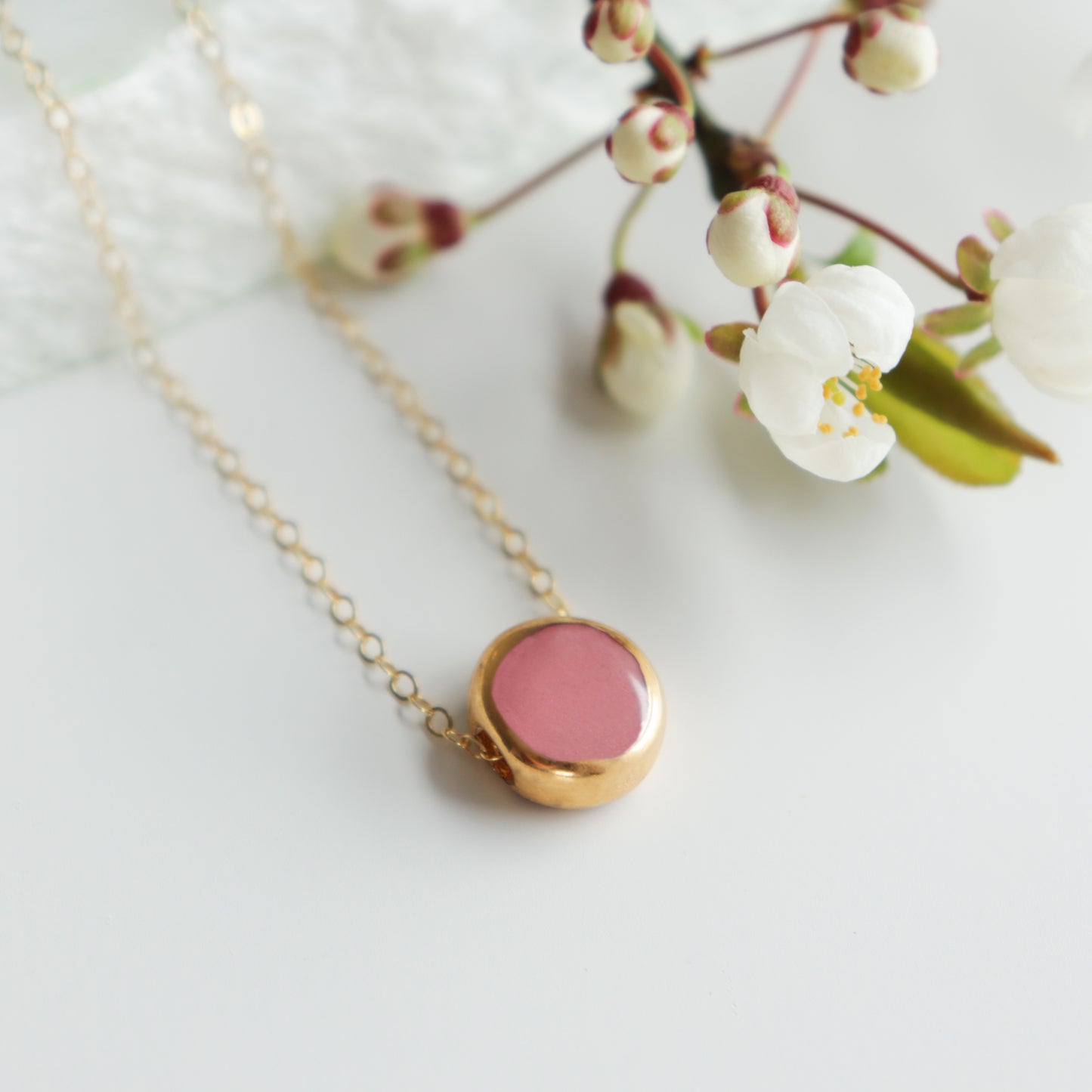 The Round Necklace by Rozenthal Jewelry, featuring a gold chain and pink round pendant in minimalist style, is displayed on a white surface beside a branch of white cherry blossom flowers.