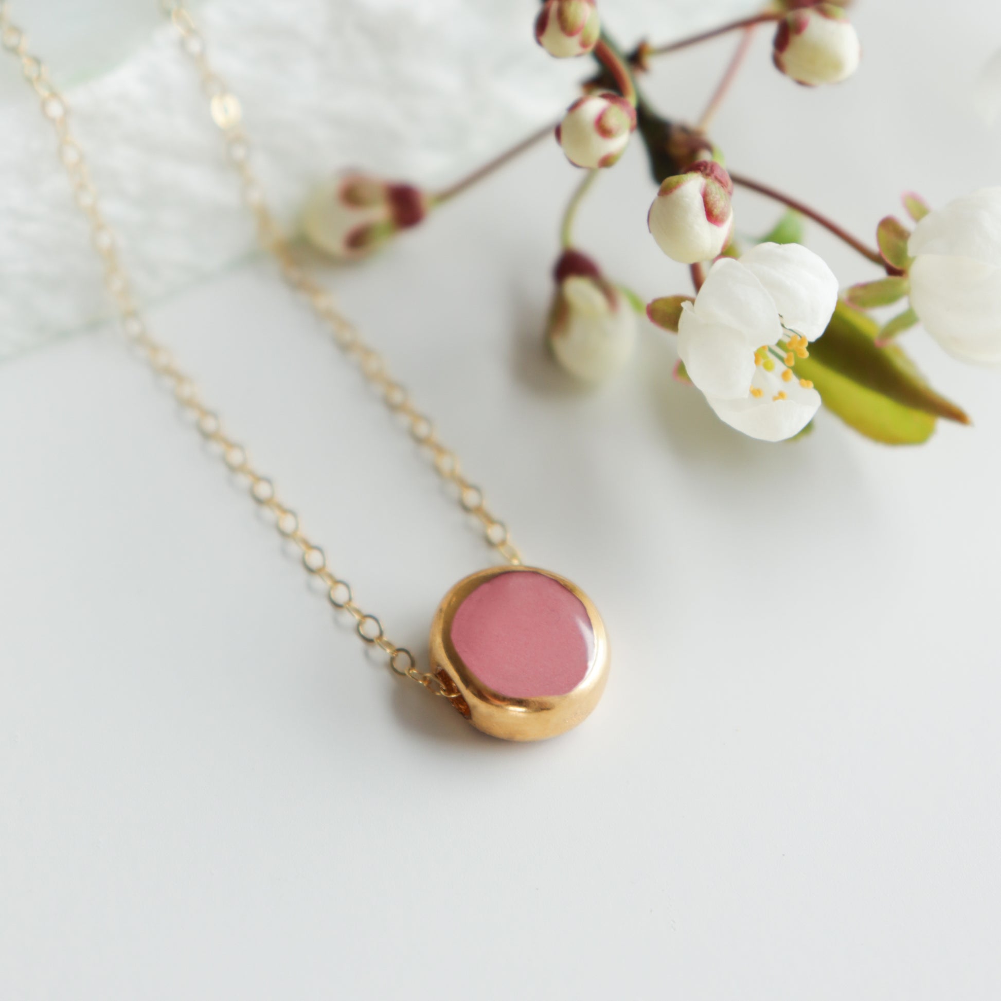 The Round Necklace by Rozenthal Jewelry, featuring a gold chain and pink round pendant in minimalist style, is displayed on a white surface beside a branch of white cherry blossom flowers.