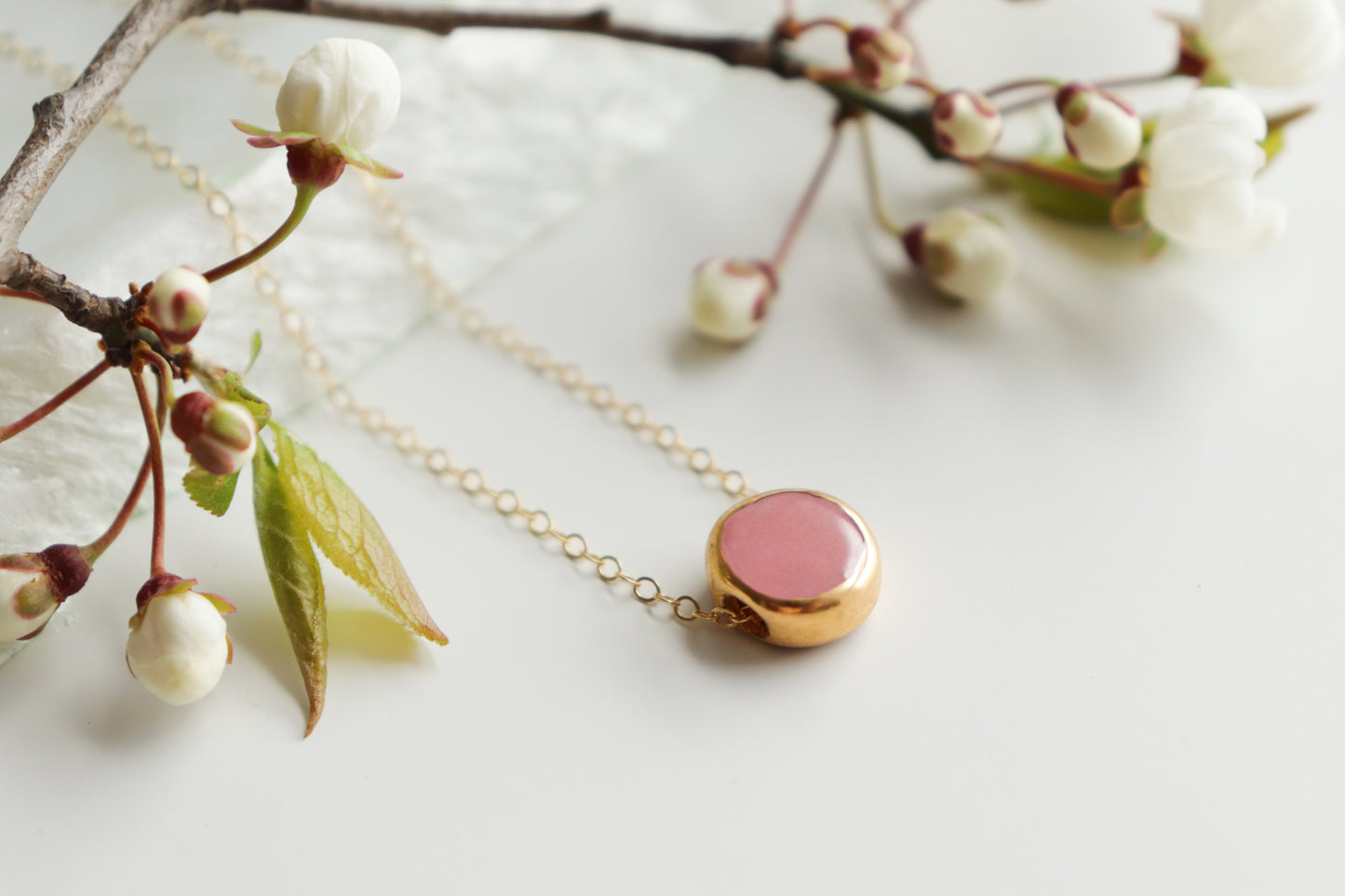 The Rozenthal Jewelry Round Necklace, featuring a round pink pendant in minimalist style, is shown on a white surface beside blooming cherry blossom branches.