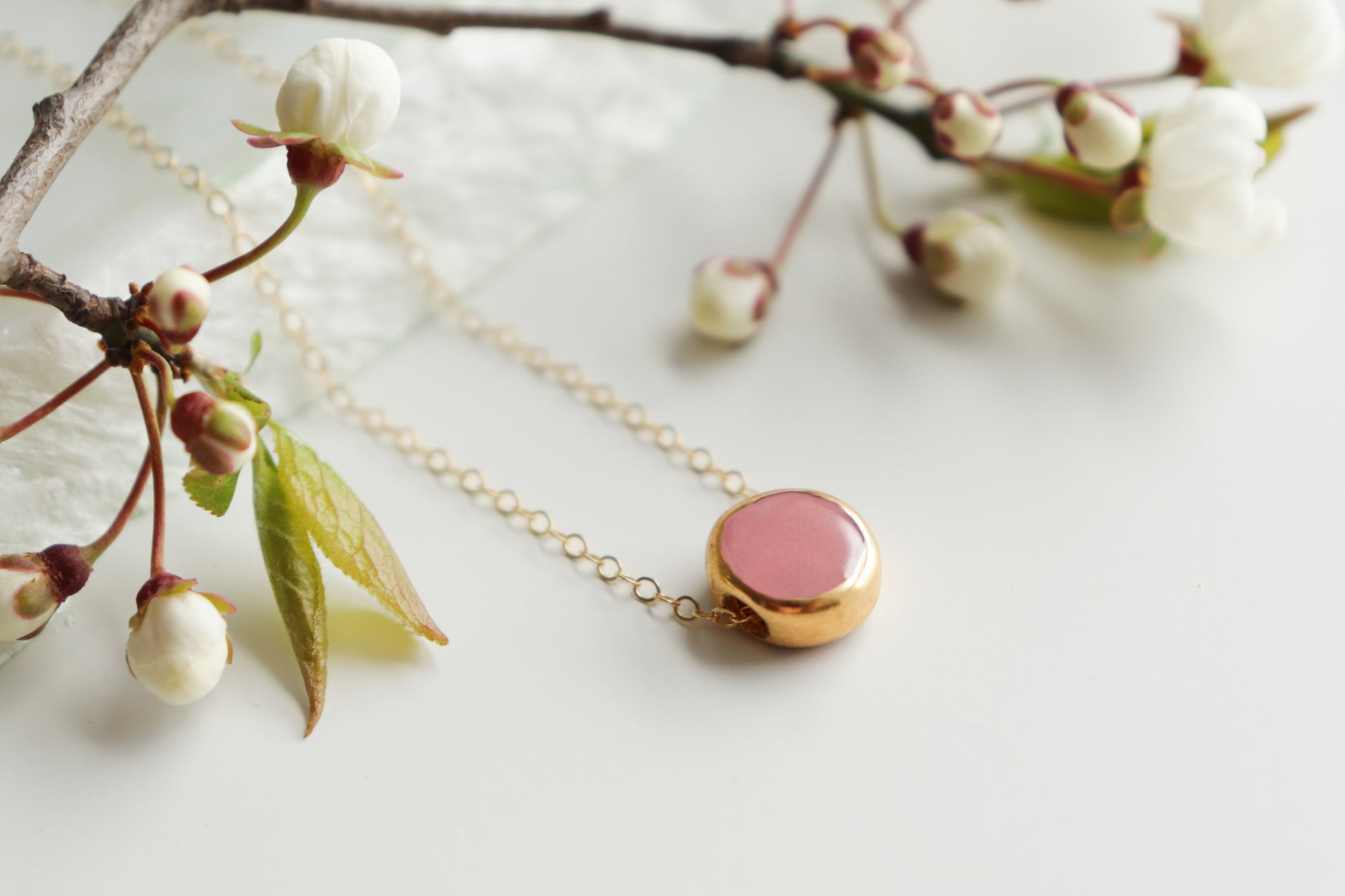 The Rozenthal Jewelry Round Necklace, featuring a round pink pendant in minimalist style, is shown on a white surface beside blooming cherry blossom branches.