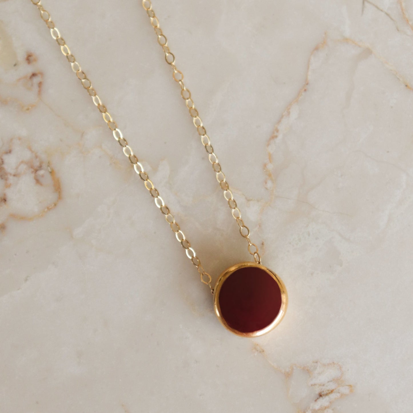 The Rozenthal Jewelry Round Necklace features a gold chain with a deep red centered round pendant, showcased on light marble. Ideal for those who love minimalist jewelry with timeless style.