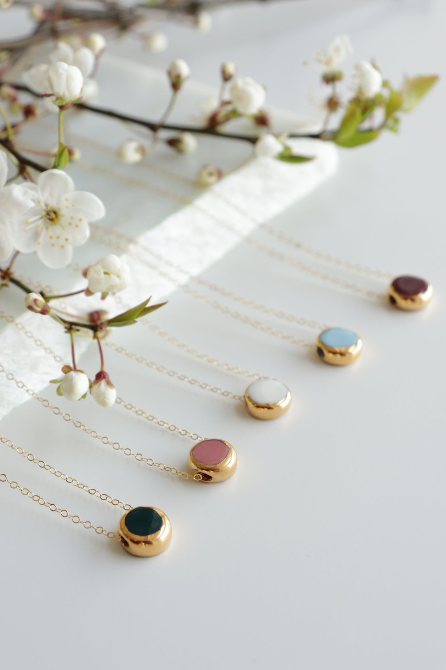 Five Rozenthal Jewelry Round Necklaces with vibrant stone pendants are arranged neatly on a white surface next to delicate white flowers, showcasing minimalist elegance in various colors.