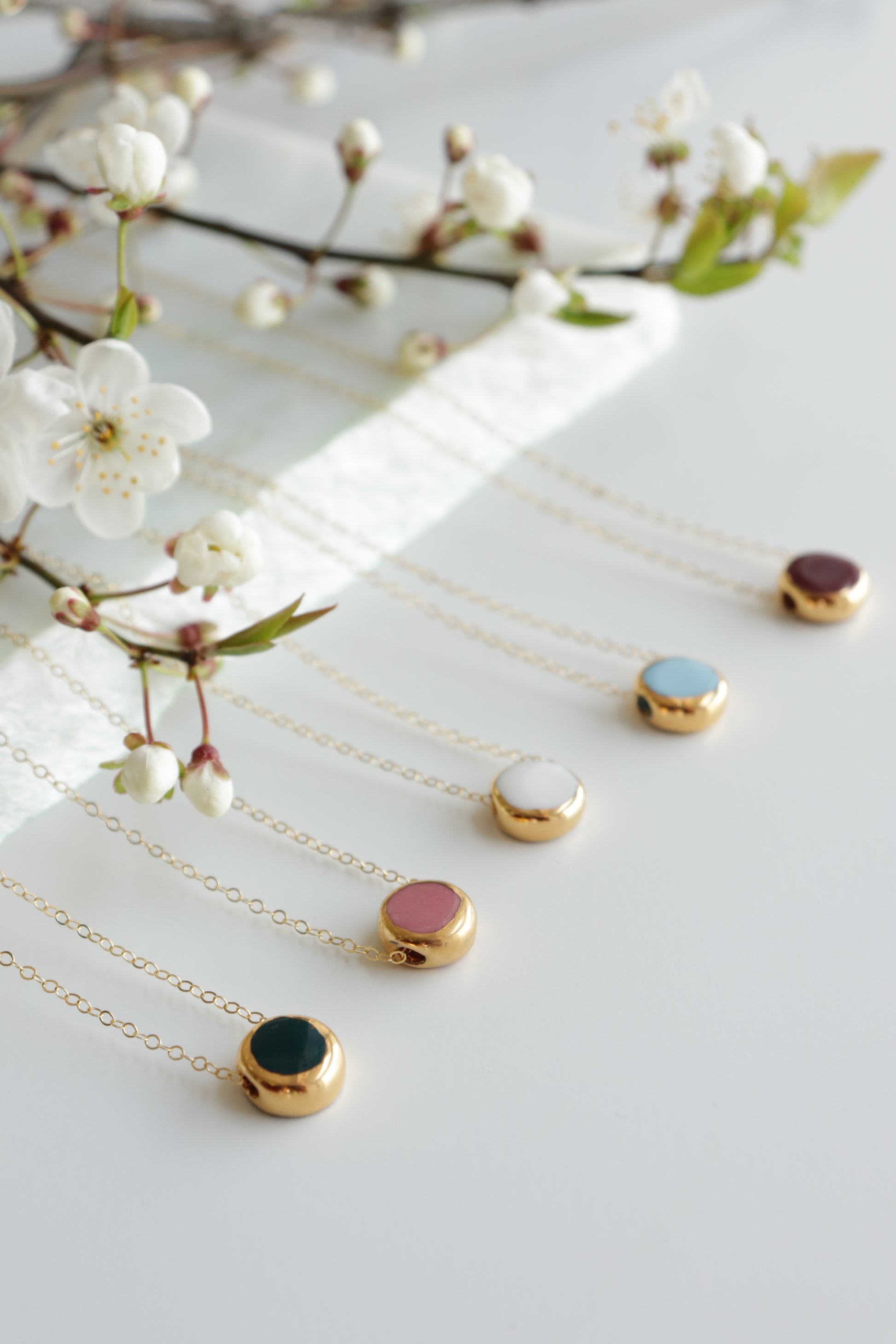 Five Rozenthal Jewelry Round Necklaces with colored pendants are arranged in a row on a white surface, accented by delicate white flowers and green foliage—a refined, minimalist showcase of elegant jewelry.