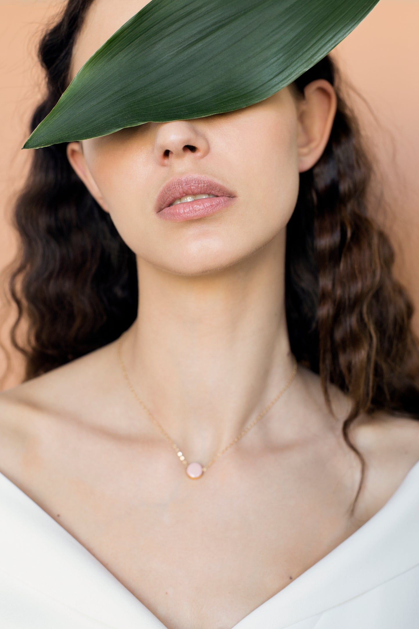 A woman with wavy brown hair wears a white top and the Rozenthal Jewelry Round Necklace, its gold gleaming above her collarbone. A large green leaf covers her eyes and forehead against a peach-colored background.