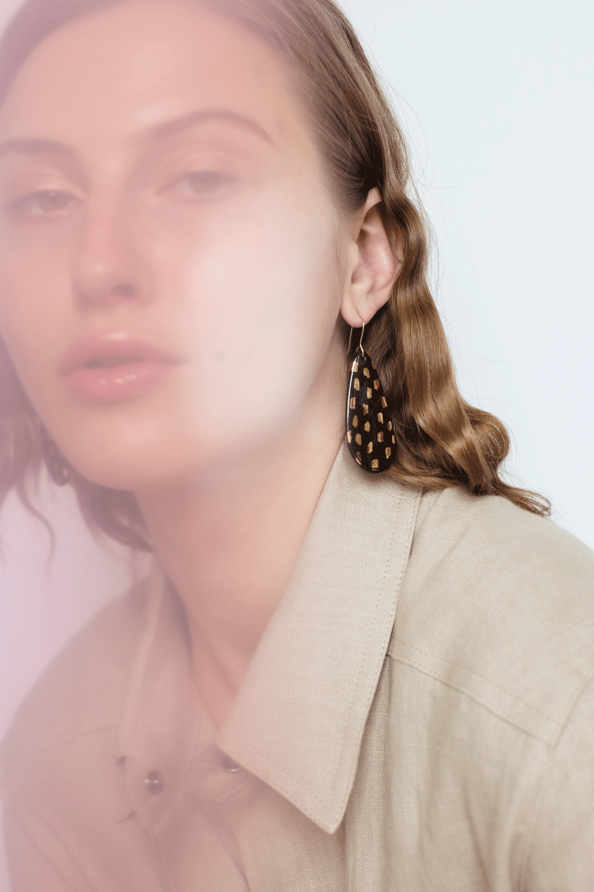 A woman with wavy light brown hair wears a beige collared shirt and Rozenthal Jewelrys Drop Earrings / L—large, handmade porcelain earrings in black and gold. The photo is softly lit with a slight pink haze.