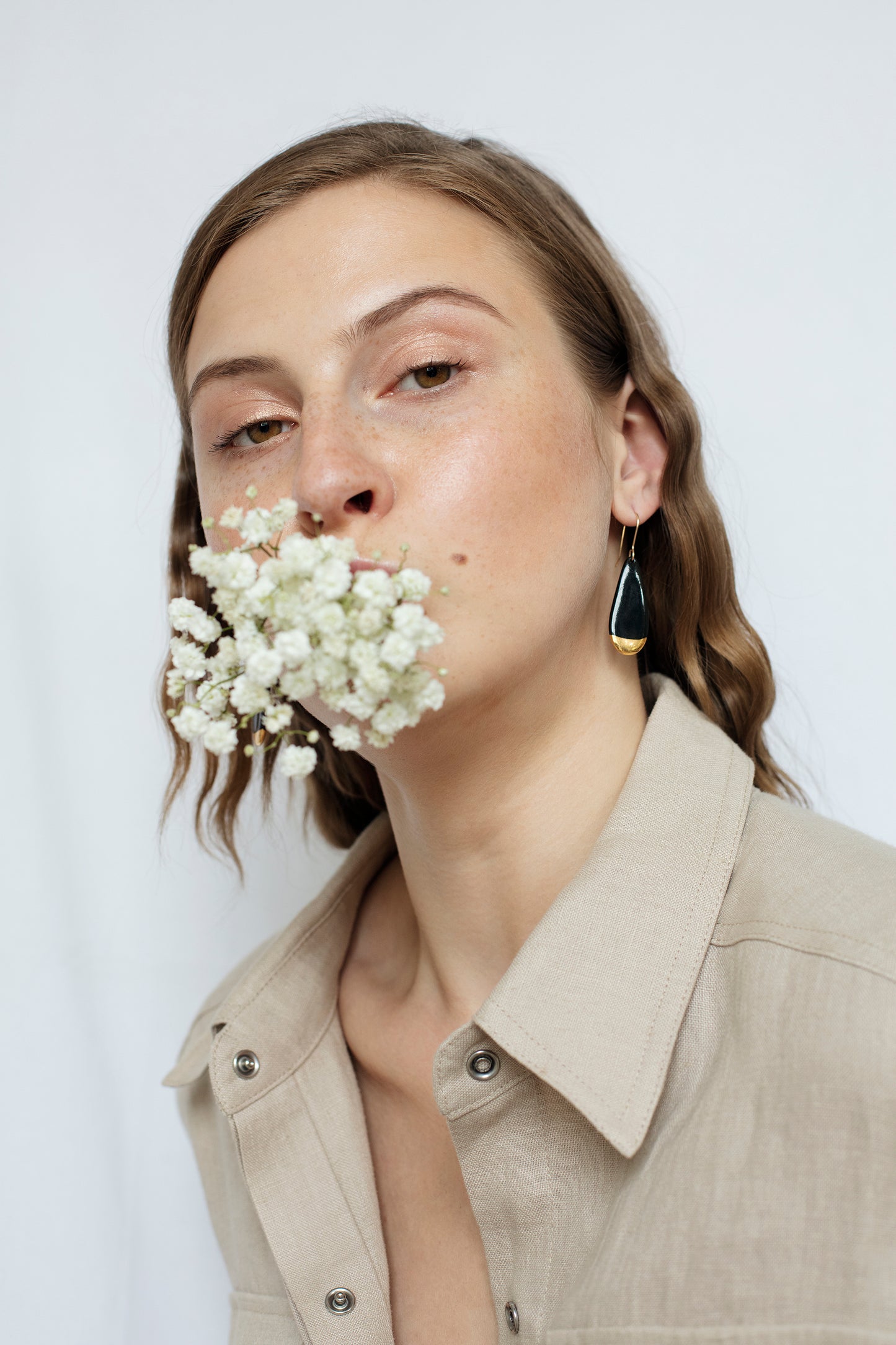 A woman with wavy brown hair wearing a beige shirt and Rozenthal Jewelry Drop earrings / S holds small white flowers in her mouth against a plain white background.
