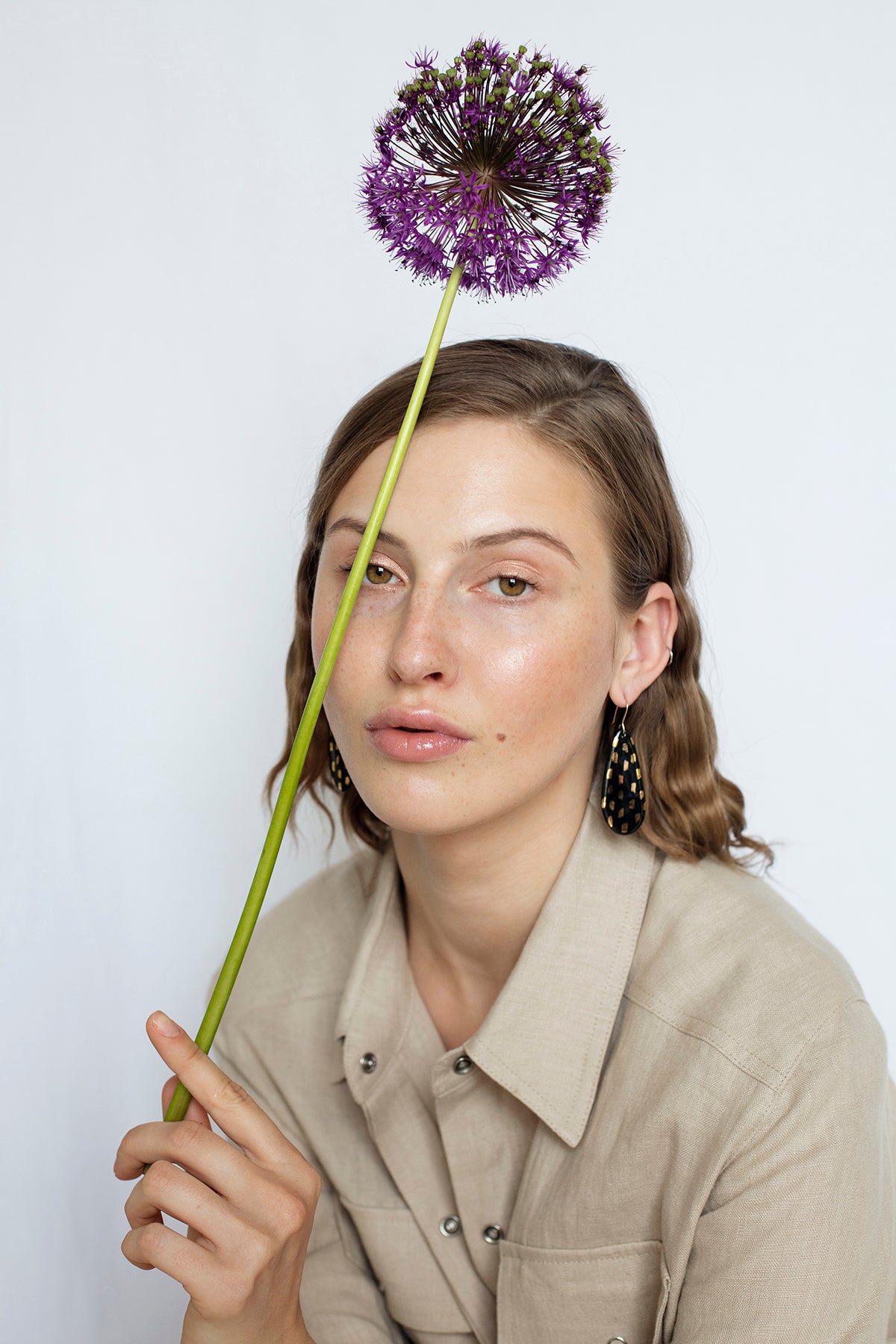 A woman with wavy brown hair and a beige shirt holds a tall purple allium before her forehead, set against a white background, embodying the elegance of Rozenthal Jewelry’s Drop Earrings / L.