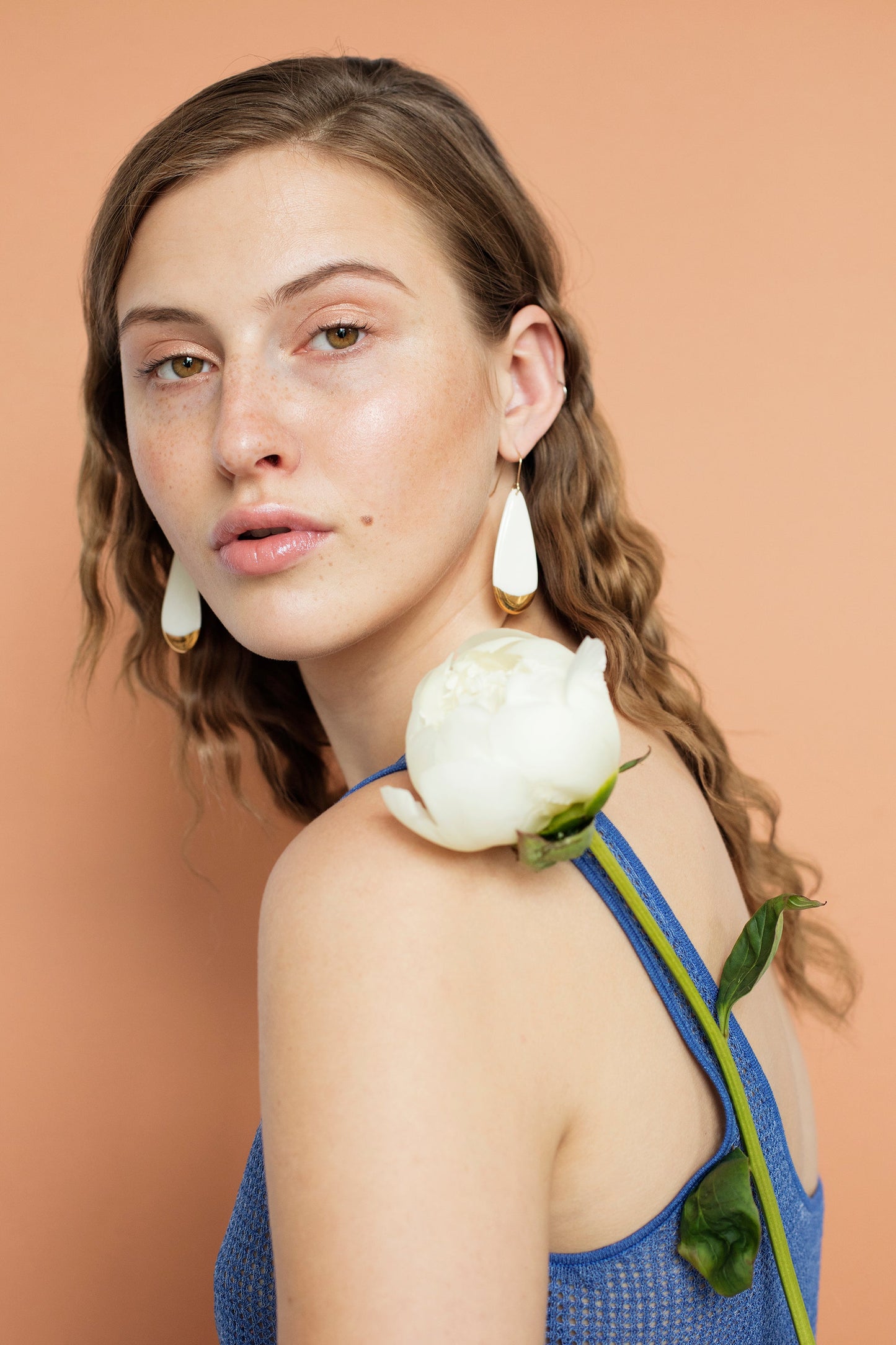 A woman with wavy light brown hair and porcelain skin wears Rozenthal Jewelrys Drop Earrings / L and a blue top, with a white peony on her shoulder, posing against a peach background.