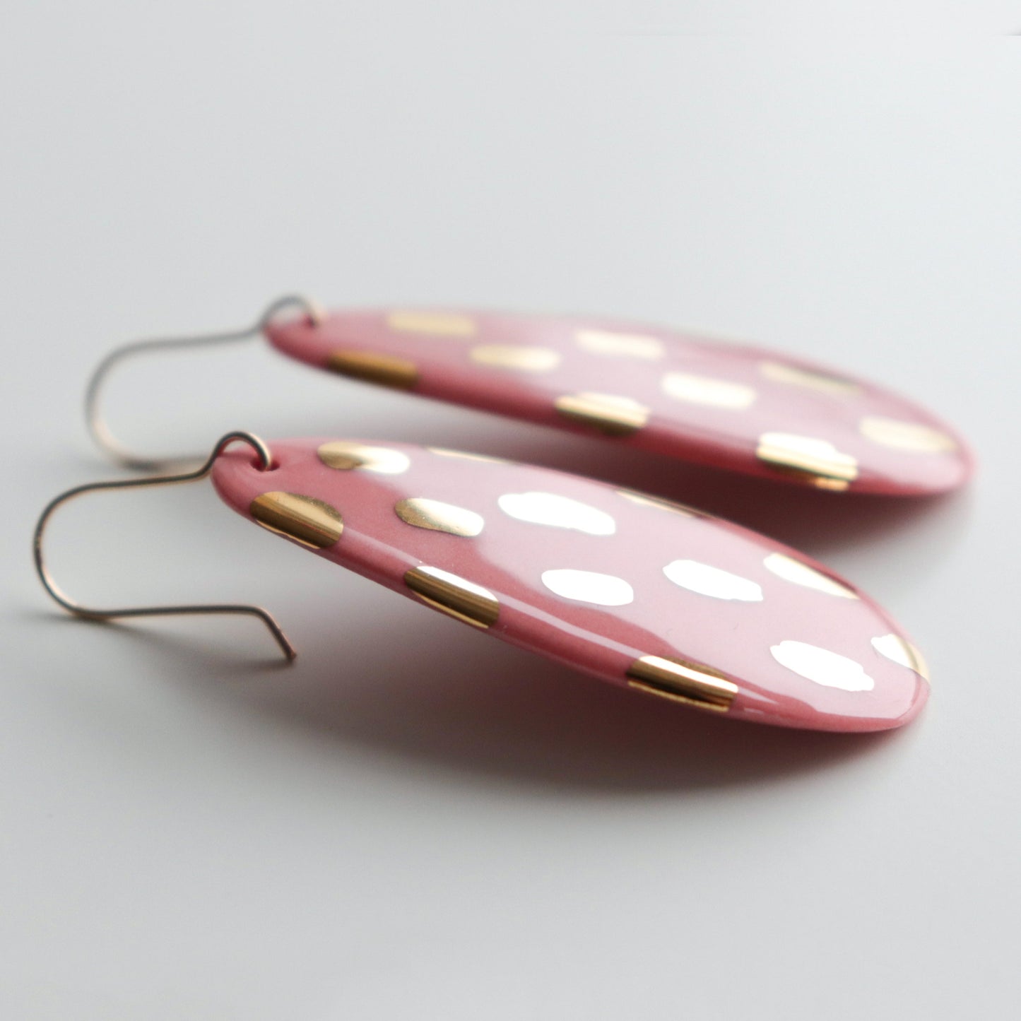 A pair of Drop Earrings / L by Rozenthal Jewelry, featuring pink oval-shaped porcelain with white spots, gold accents, and minimalist metal hooks, displayed on a white surface.