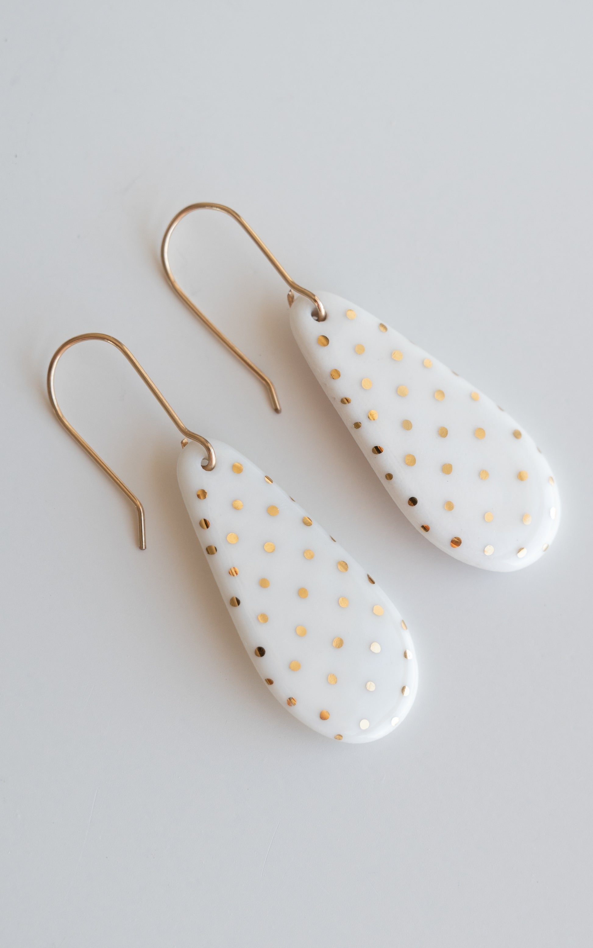 Drop earrings / S by Rozenthal Jewelry: teardrop-shaped white porcelain statement earrings with gold polka dots and hooks, handmade in Latvia and shown on a light gray background.