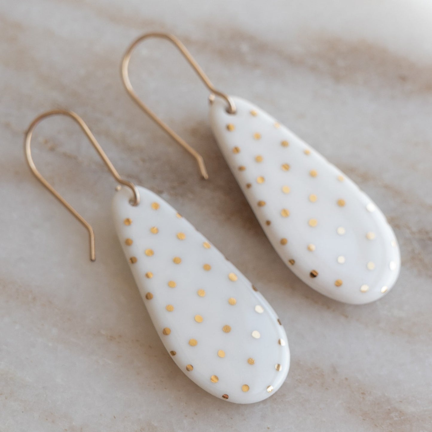 Drop earrings / S by Rozenthal Jewelry—handmade in Latvia, featuring teardrop-shaped white ceramic with gold polka dots and gold hooks. Unique, sustainable jewelry resting on beige marble—perfect for any occasion.