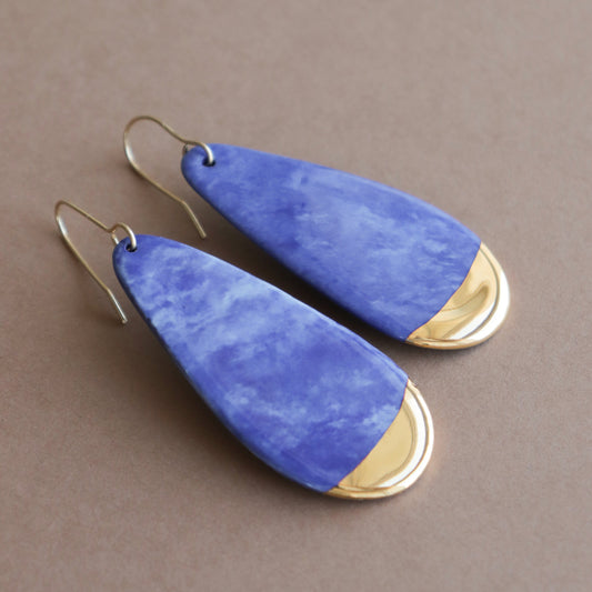 The Drop Earrings / L by Rozenthal Jewelry are teardrop-shaped porcelain earrings with a marbled blue and white design and gold accents—ideal for minimalist style. Shown against a neutral brown background.
