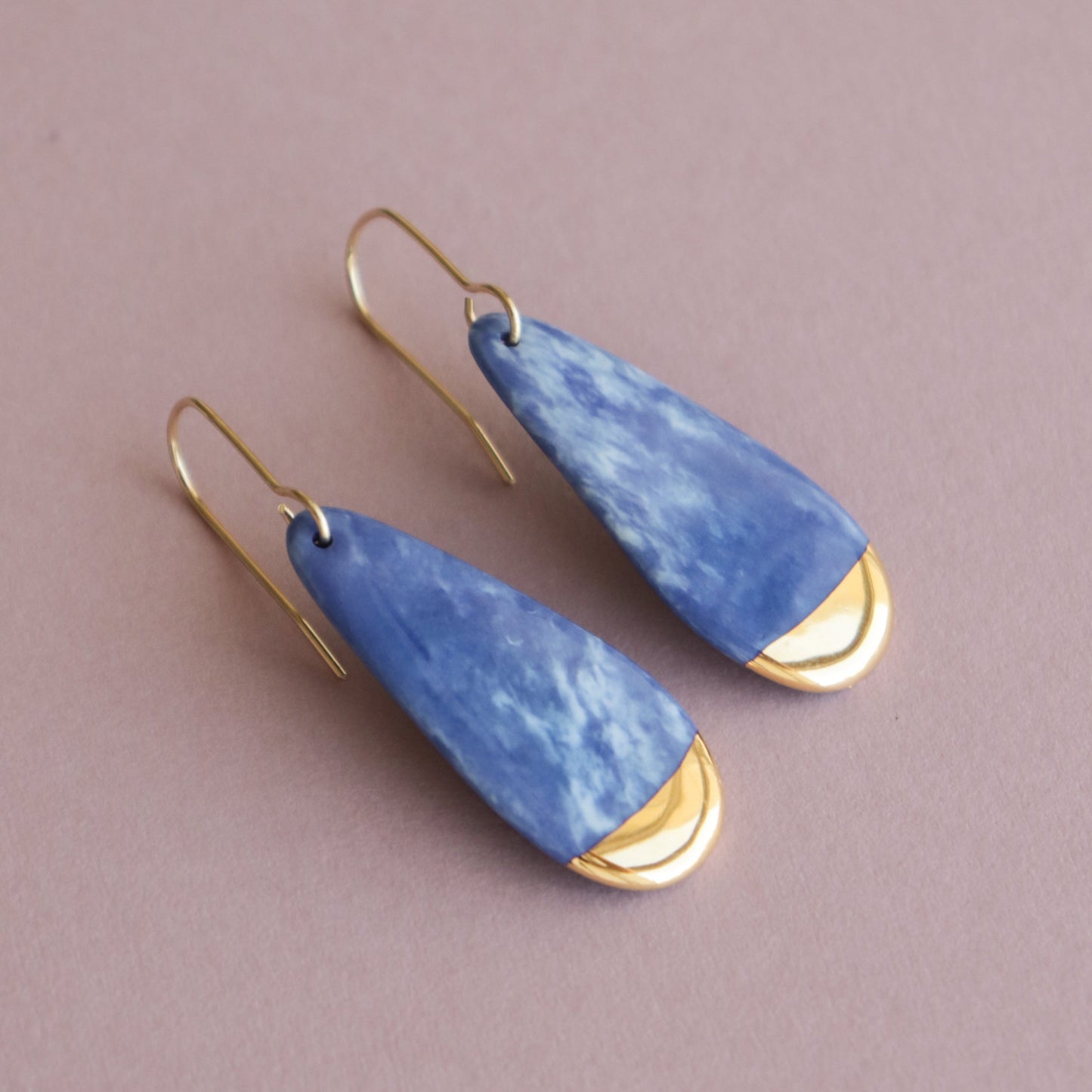 Rozenthal Jewelrys Drop earrings / S are teardrop-shaped porcelain statement earrings with blue marbled stone and gold accents, handmade in Latvia and shown on a light mauve background.