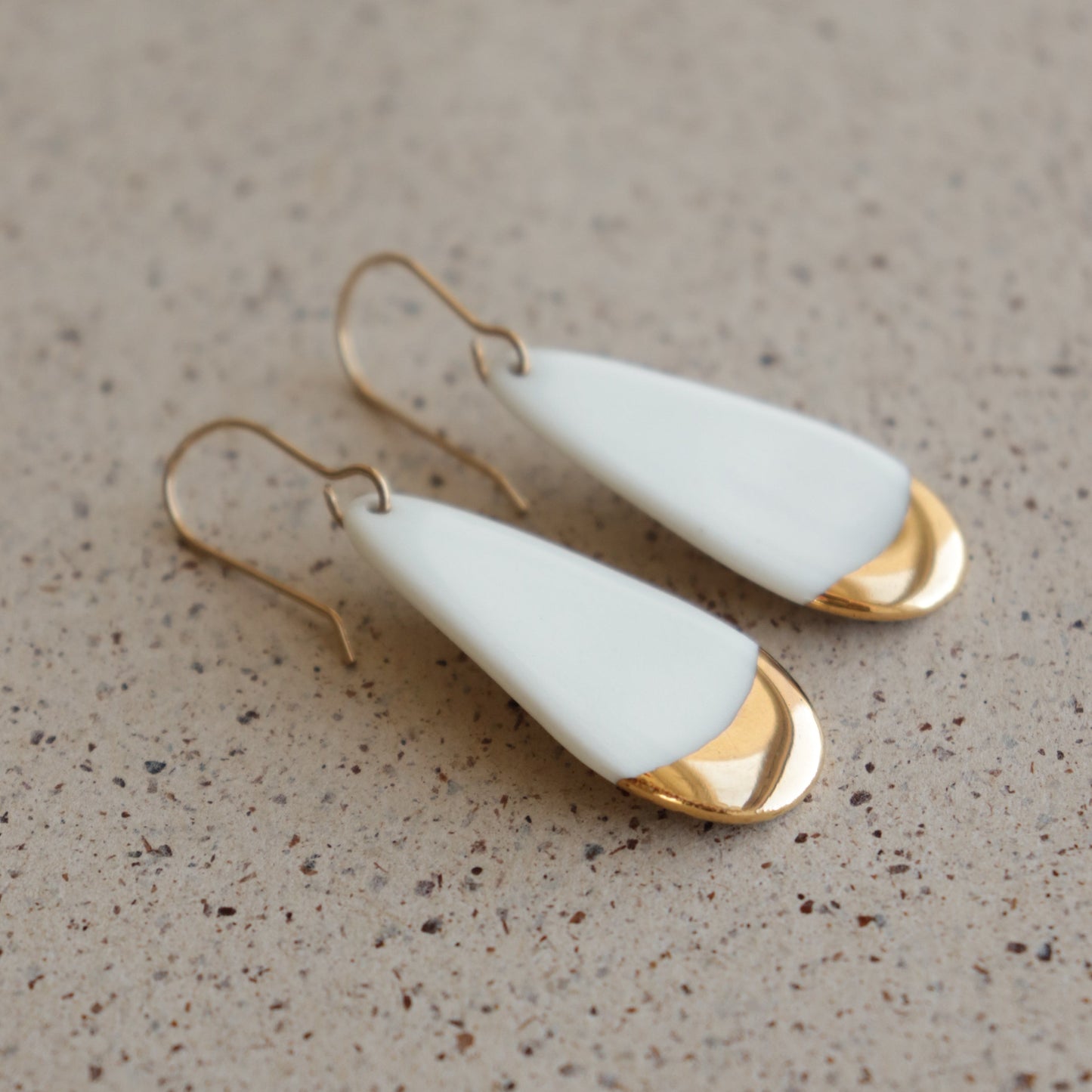 Drop earrings / S by Rozenthal Jewelry: handmade in Latvia, these teardrop-shaped porcelain statement earrings feature a smooth white top, shiny gold accents at the base, and gold hooks. Pictured on a speckled beige surface.