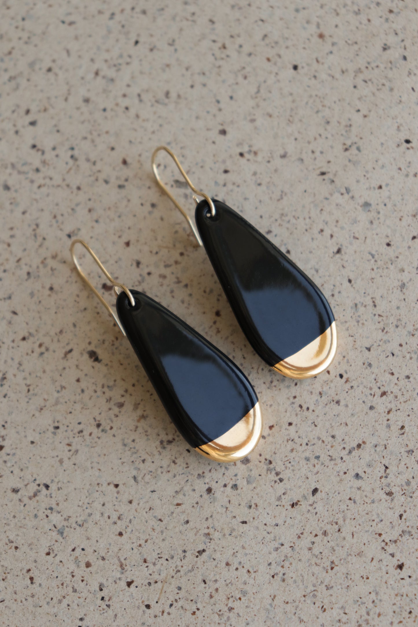 Rozenthal Jewelry’s Drop earrings / S feature teardrop-shaped porcelain with glossy black surfaces and gold accents. Lightweight, waterproof, and chic, they are displayed on a speckled beige surface—perfect for elevating any look.