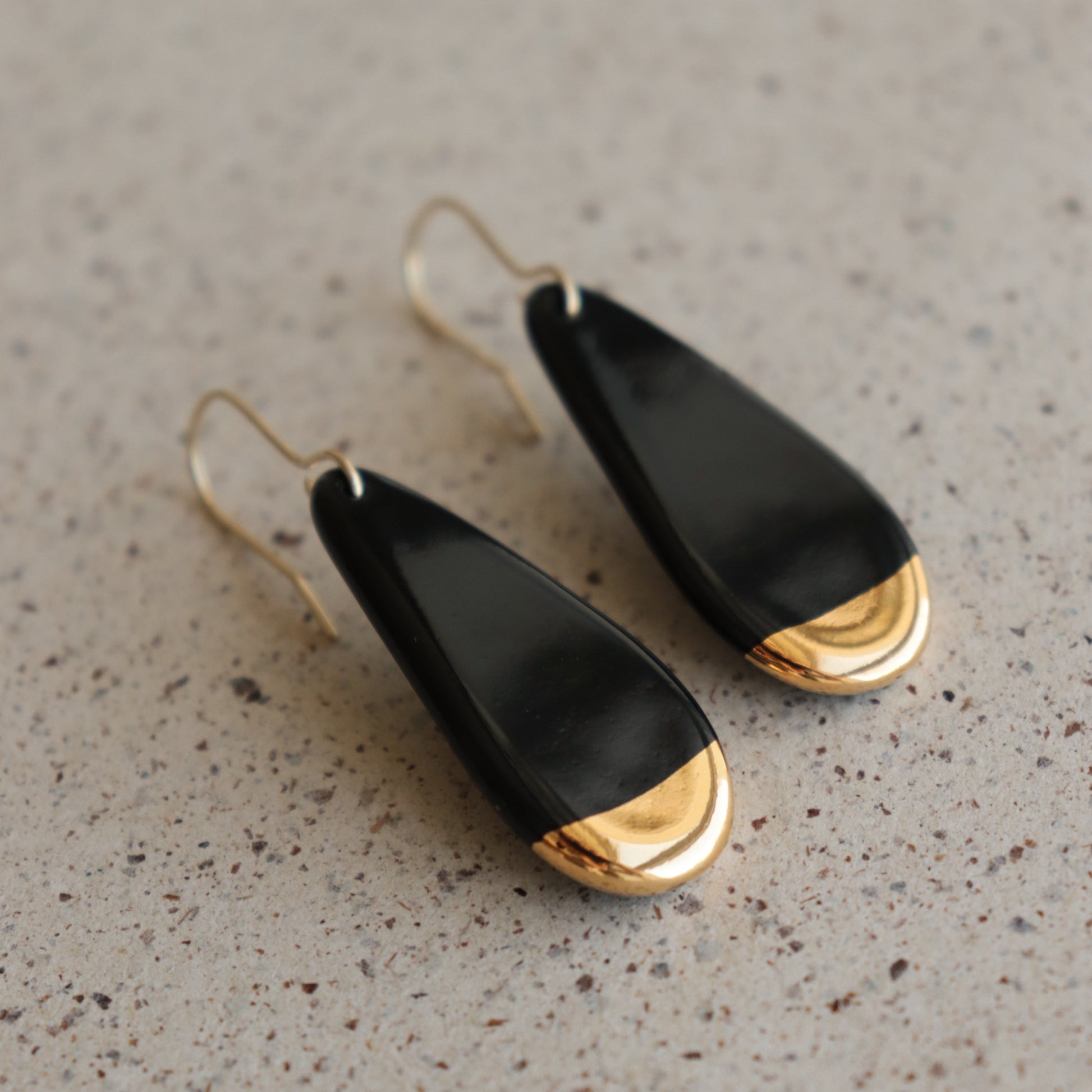 The Rozenthal Jewelry Drop earrings / S are teardrop-shaped, handmade in Latvia with black bodies, gold tips, and gold hooks. Lightweight and waterproof, they rest stylishly on a speckled beige surface.